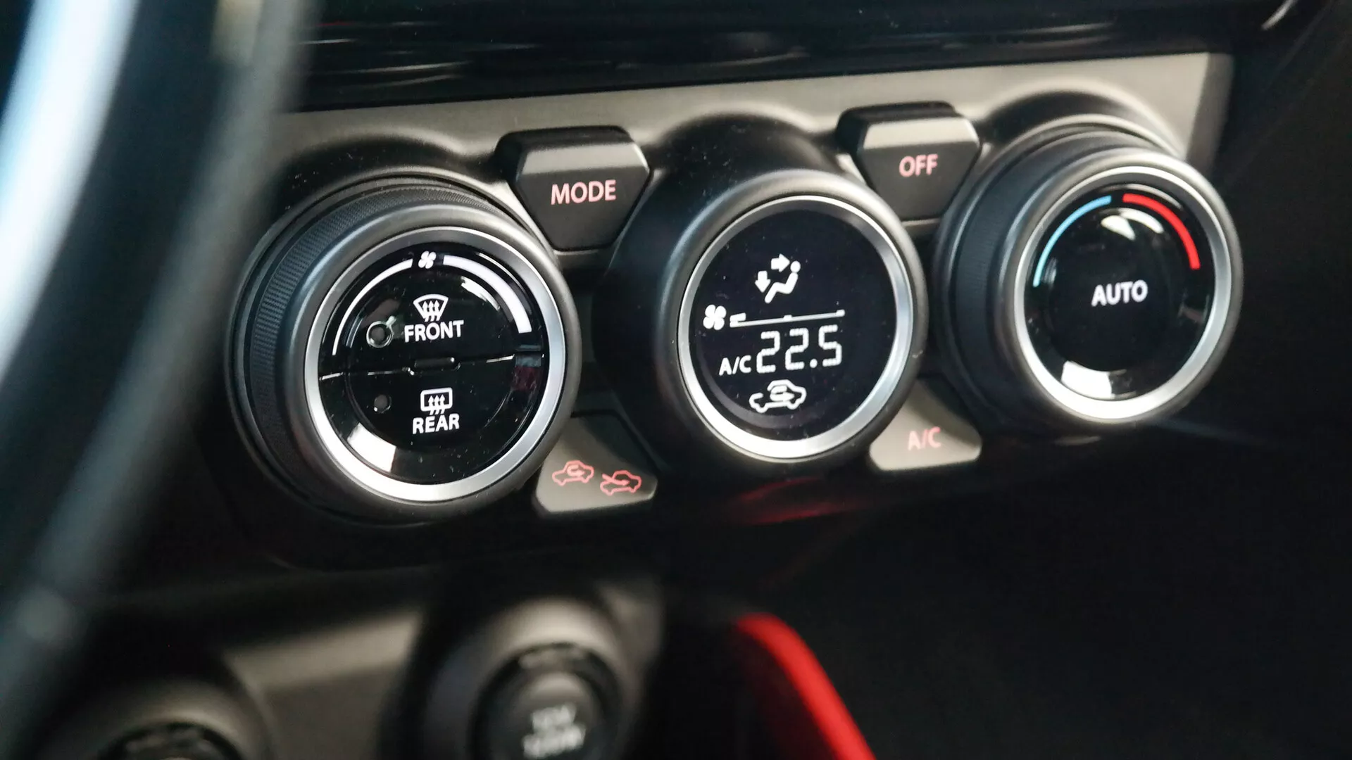 Povečani pogled na klimatski sistem Suzuki Swift Sport 2017 iz perspektive vozniškega sedeža. Slika poudarja sredinsko konzolo s tremi črnimi krožnimi gumbi z belimi in rdečimi poudarki; levi gumb označen 'MODE' nadzoruje smer pretoka zraka, srednji gumb prilagaja moč in hitrost ventilatorja s položajem 'OFF', desni gumb pa prikazuje digitalno temperaturo 22,5 stopinj Celzija, kar pomeni avtomatski klimatski sistem s funkcijami 'AUTO' in 'AC'. Pod temi gumbi so delno vidni dodatni gumbi, vključno z gumbom s simbolom za odtaljevanje. Ozadje je zamegljeno, da poudari kontrole klimatskega sistema.