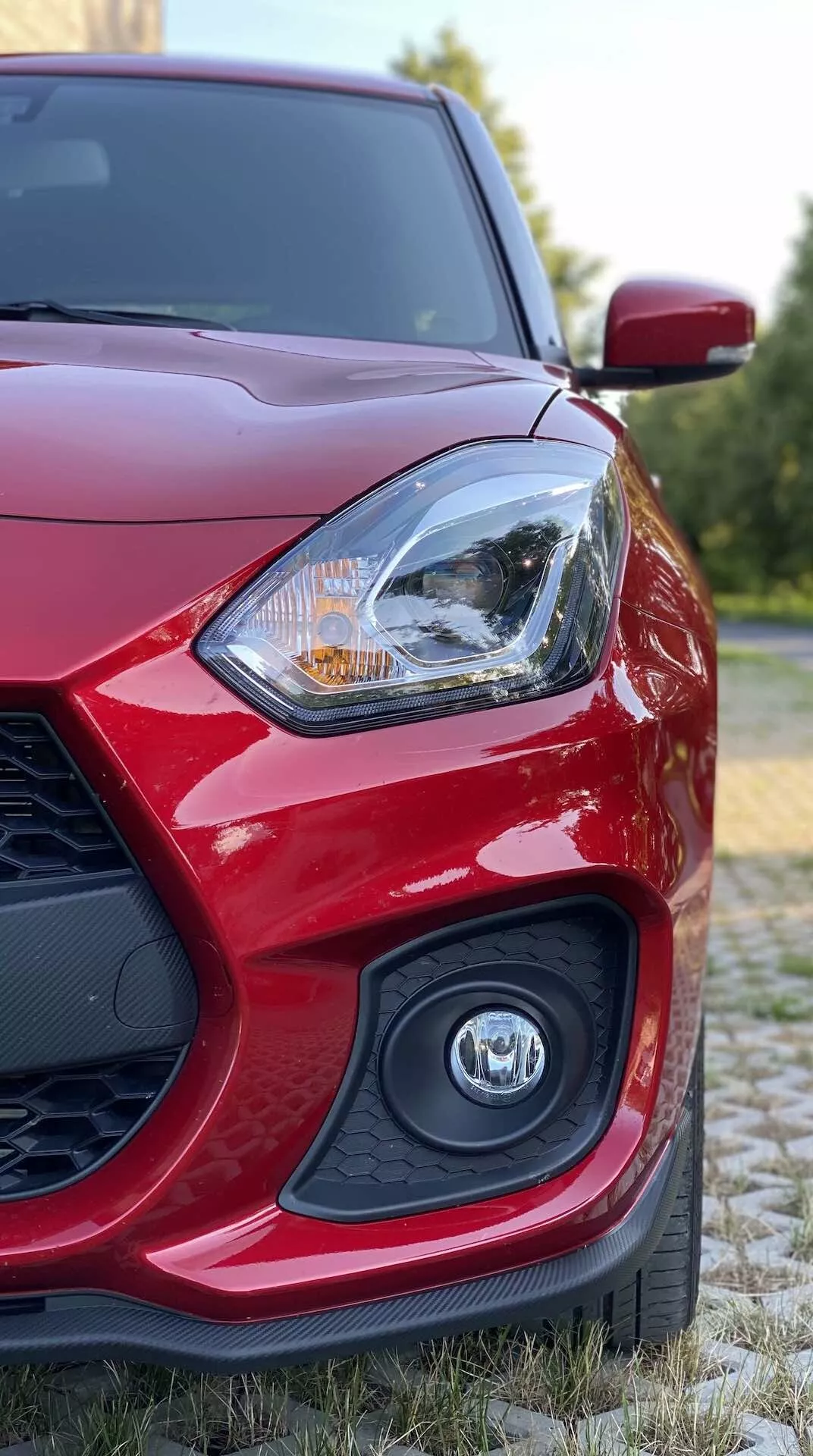 Pogled od blizu na rdeči Suzuki Swift Sport 2017 z nizkega kota, osredotočen na sprednji levi del, ki prikazuje voznikovo luč z zapletenim dizajnom, ki lahko vključuje LED ali halogensko tehnologijo osvetlitve, delno zamegljenim emblemom na rešetki in aerodinamično stransko ogledalo v naravnem okolju z zelenjem.