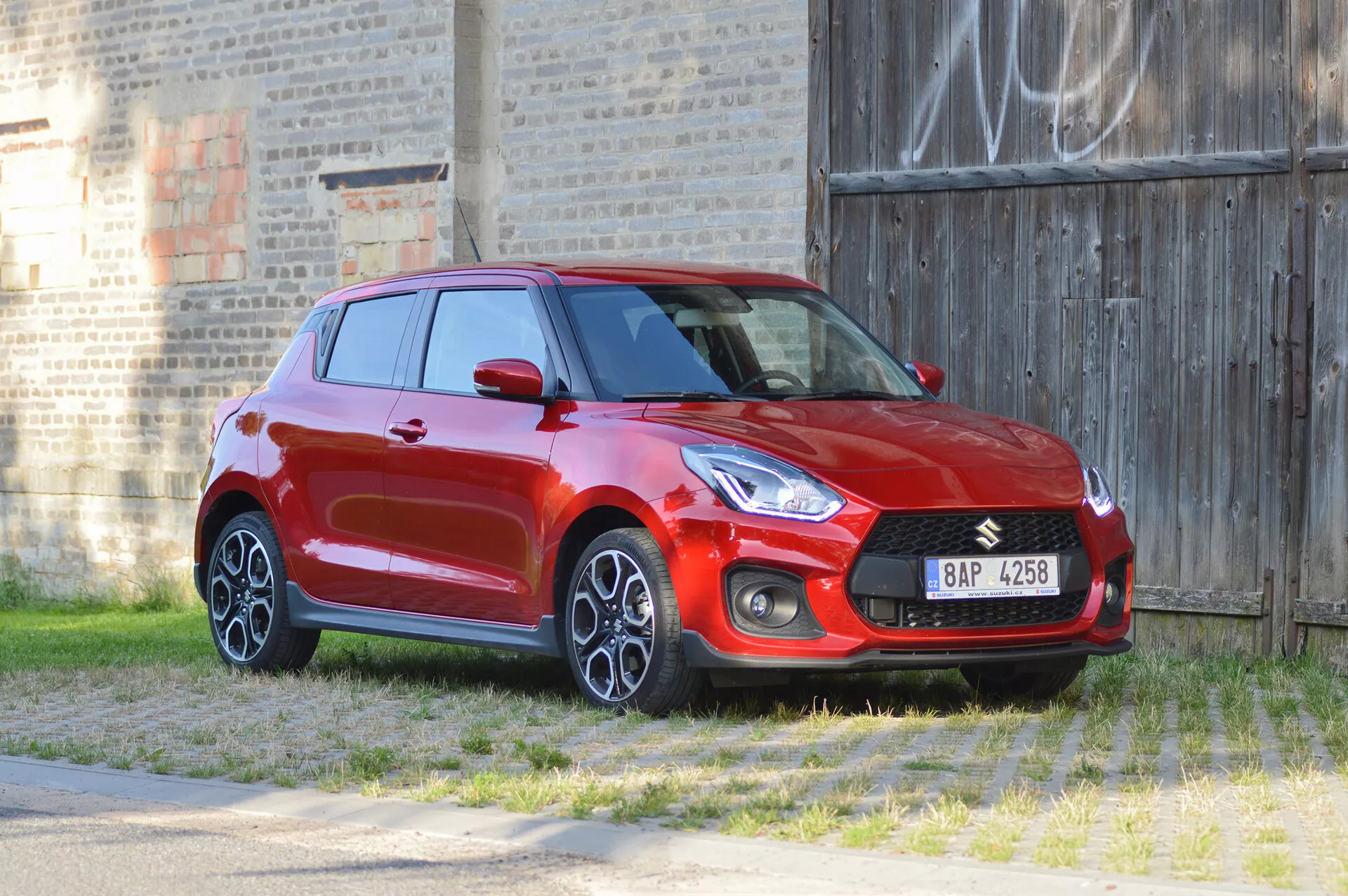 Slika rdečega hatcbacka Suzuki Swift Sport 2017, posneta iz tridčetrtinskega sprednjega kota, poudarja sprednje in stranske profile. Oblikovne značilnosti avtomobila vključujejo izrazito sprednjo masko z vgrajenim logotipom, elegantne žaromete, ohišja meglenk in večkraka lita platišča. Posnetek je narejen z rahle višine, kar omogoča pogled od zgoraj navzdol na pokrov motorja in streho. Okolica vključuje travnato ospredje in zid iz opeke z grafiti v ozadju, kar poudarja športen in dinamičen videz vozila.