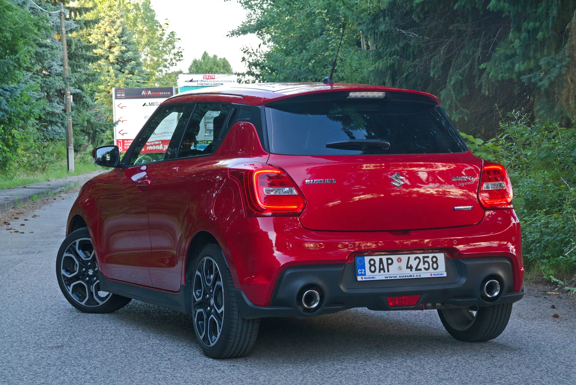Ta slika prikazuje zadnji tristranski kotni pogled na rdečo hatchback vozilo Suzuki Swift 2017 (Swift-Sport), ki zajeme zadnjo desno stran vozila. Fotografija poudarja moderen športni dizajn avtomobila z značilnimi funkcijami, kot so LED zadnje luči, zadnji brisalec vetrobranskega stekla, osrednji zadnji znak z informacijami o modelu, vidna izpušna cev, strešni spojler, večkraka aluminijasta platišča in zadnji odbijač opremljen s sistemom parkirnih senzorjev. Dobro osvetljena fotografija, posneta v naravni dnevni svetlobi, nakazuje, da je vozilo parkirano na primestni cesti obdani z drevesi, s jasno vidno streho in anteno. Registrska tablica je zamegljena za zagotovitev zasebnosti.