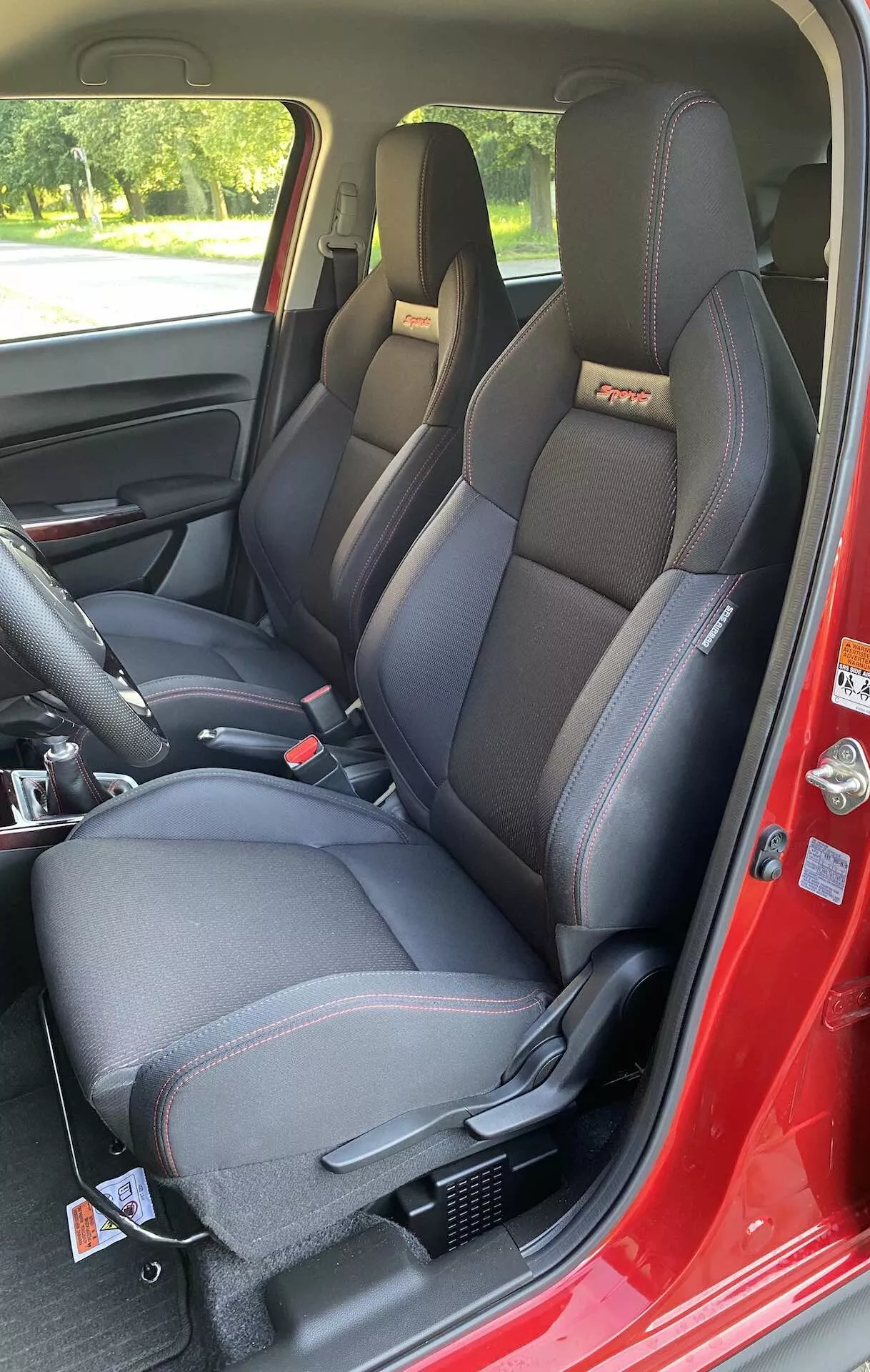 Notranjost Suzuki Swift Sport 2017 je prikazana iz perspektive sopotnika skozi odprta sprednja vrata. Osredotočamo se na moderna, športna oblikovana sprednja sedeža z vgrajenima vzglavnikoma in izrazitimi stranskimi oporami, najverjetneje v kombinaciji umetnega usnja in tkanine s kontrastnimi šivi. Na zgornjem delu naslonjal so vidne vtisnjene ali vezene logotipi. Oba sedeža sta opremljena s tritočkovnim varnostnim pasom, voznikova stran pa ima nastavljiv mehanizem za višino pasu na stebričku B. Sredinska konzola med sedeži ima shranjevalne predale in ročico ročne zavore. Notranji temen panel vrat vključuje kljuko, nadzor za prilagajanje ogledal in električne kontrole oken. Tla so prekrita s temno preprogo, osnova sedežev pa prikazuje pritrdilne letve. Dnevna svetloba in naravno zunanje okolje sta vidna skozi okna, z odboji in sencami, ki poudarjajo podrobnosti kabine.