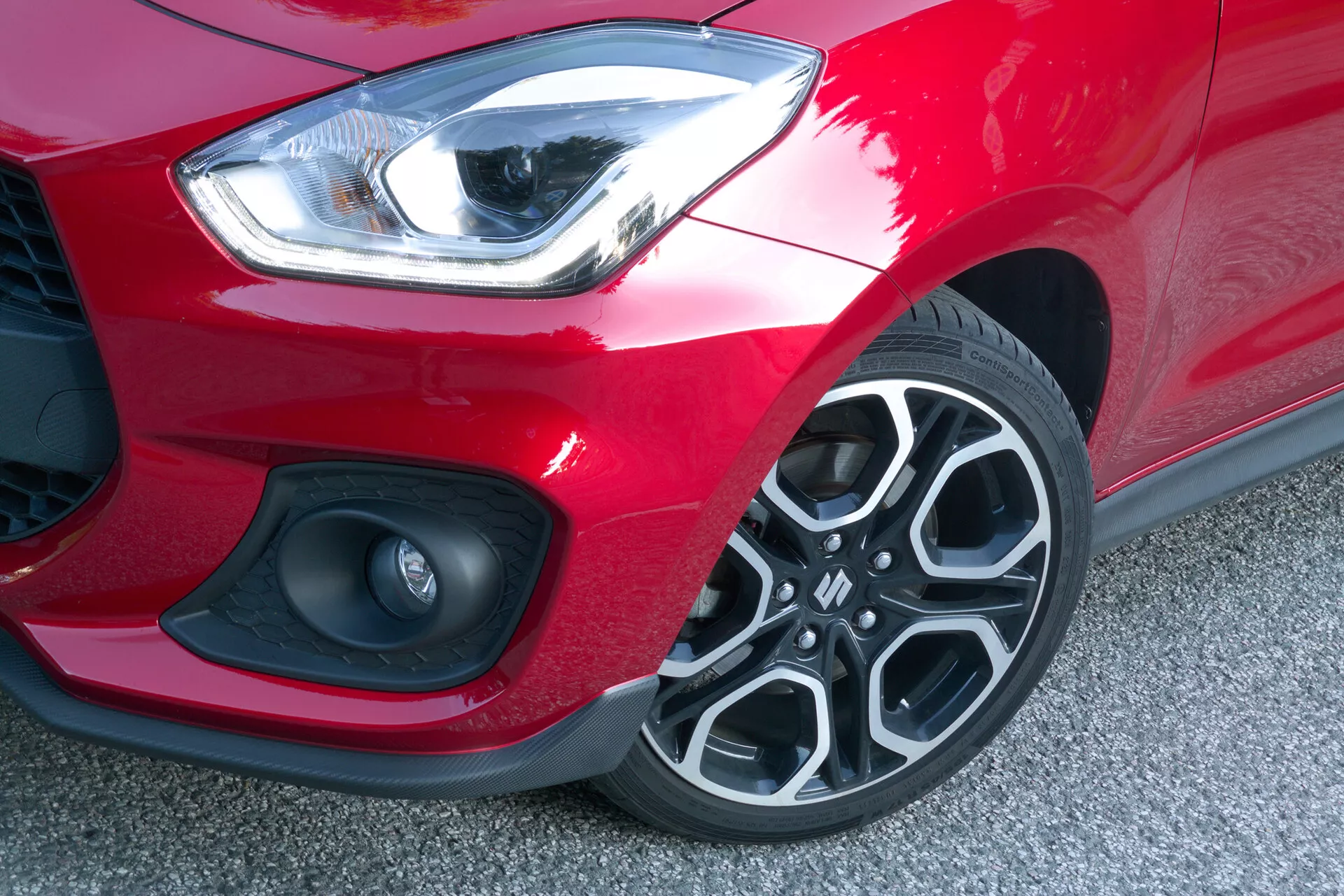 Podrobna slika, ki prikazuje prednji levi kot Suzuki Swift Sport 2017 iz vogalnega in rahlo sprednjega kota. Ukrivljen in eleganten LED žaromet oddaja močno belo svetlobo, kar kaže na funkcijo dnevne luči. Pod žarometom je videti majhen del mrežaste rešetke, poleg krožnega elementa, ki bi lahko bil meglenka ali parkirni senzor, nameščenega v dinamično oblikovanem sprednjem odbijaču. Mat črn del odbijača je v kontrastu s svetlo rdečo barvo avtomobila. Večkraki dizajn platišč iz lahke zlitine s temnimi in srebrnimi elementi izstopa, z vidnim profilom pnevmatike, kar kaže na dobro stanje. Nizka višina avtomobila od tal je opazna, kar odraža standardno višino, primerno za športni model.