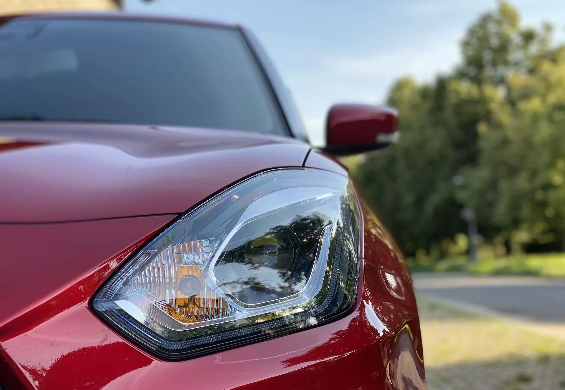 Povečava sprednjega levega žarometa avtomobila Suzuki Swift Sport 2017. Slika je posneta iz sprednje tretje perspektive, poudarja oblikovne podrobnosti žarometa z jasno lečo in zapletenim notranjim reflektorjem ter oranžno smerno lučjo. Vidni so sijoči, globoko rdeči karoserijski paneli, vključno s pokrovom motorja in blatnikom, na zamegljenem zunanjem ozadju z drevesi in svetlim nebom.