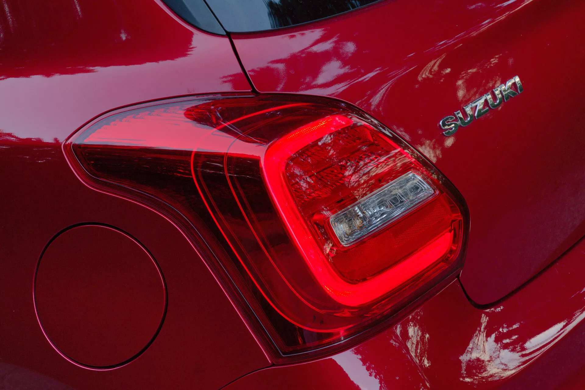 Povečan posnetek zadnje leve luči avtomobila Suzuki Swift Sport 2017, ki poudarja segmentiran dizajn svetlobnih enot vključno z zavorno lučjo in smerno svetilko ali vzvratno lučjo. Karoserija v globoki rdeči barvi s sijajnim premazom odseva svetlobo in okolico. Poleg zadnje luči je viden kromiran emblem z napisom 'SWIFT'. Slika prav tako razkriva okrogel pokrov za gorivo in prikazuje moderen jezik oblikovanja z aerodinamičnimi linijami in trpežnimi materiali za konstrukcijo.