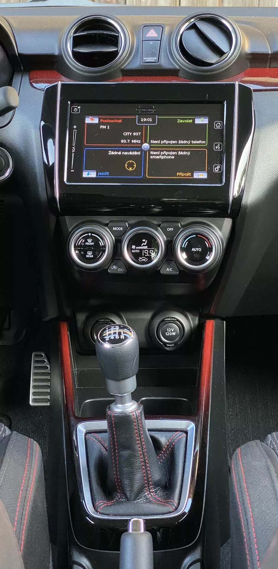 Sredinska konzola v notranjosti Suzuki Swift Sport 2017, z infotainment sistemom s fizičnimi gumbi, okroglimi zračniki, ročnim upravljanjem klimatske naprave s tremi gumbi za nastavitev temperature, hitrosti ventilatorja in razporeditve zraka ter športno prestavno ročico. Konzola vključuje usnjeno oblogo, okrasne elemente z vzorcem iz ogljikovih vlaken in športno rdečo črto. Dodatni nadzorni elementi pod prestavno ročico kažejo na napredne sisteme za pomoč pri vožnji.