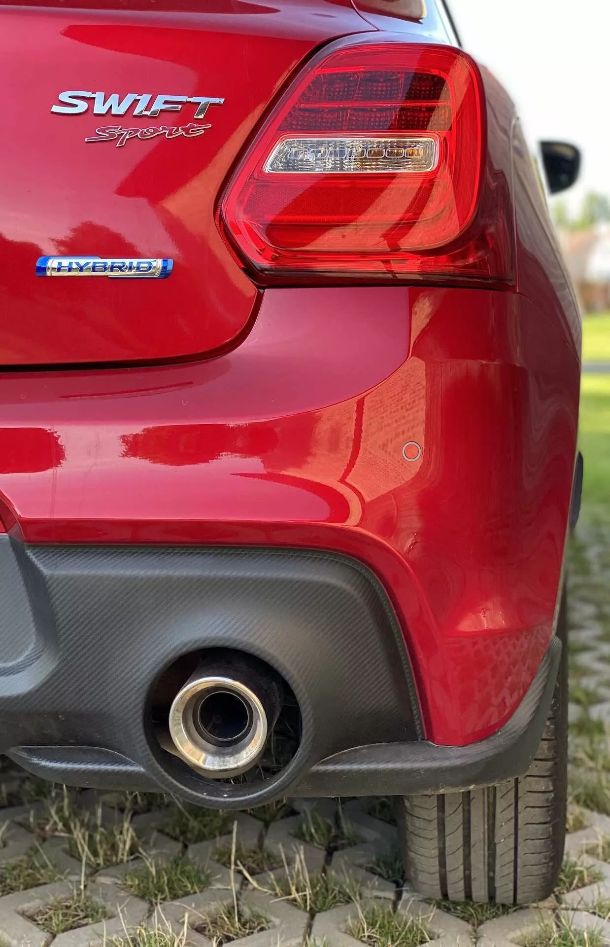 Podroben pogled zadnjega dela rdečega hatchbacka Suzuki Swift 2017 iz nizkega kota. Slika se osredotoča na desno zadnjo luč s prozorno lečo in rdečimi osvetljevalnimi sekcijami, značko 'SWIFT' pod lučjo, značko 'HYBRID' spodaj, kar označuje hibridni pogon, delno zamegljen zadnji del, desno zadnjo pnevmatiko z vidnim vzorcem tekalne plasti, črni kontrastni del na zadnjem odbijaču z morebitnim aerodinamičnim vstopom in okroglo kovinsko izpušno cev v spodnjem desnem kotu, ki odbija svetlobo.