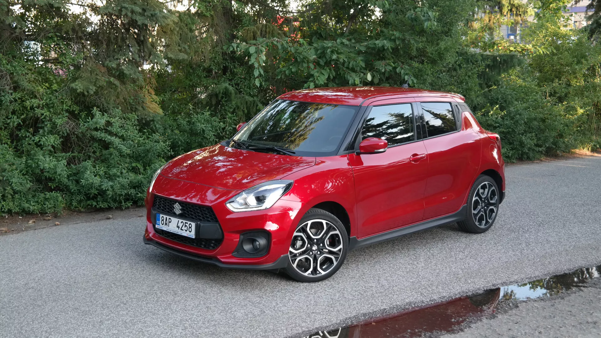 Slika rdečega 2017 Suzuki Swift Sport hatchbacka, posneta iz treh četrtinskih sprednjih kotov, ki prikazuje elegantne žaromete, izrazito prednjo masko z vodoravnimi črtami, gladek pokrov motorja, jasen vetrobran, voznikovo stran z zrcalom, barvo telesa, večkraka platišča iz zlitine in strešni spojler proti tlakovani cesti z naravnim okoljem.