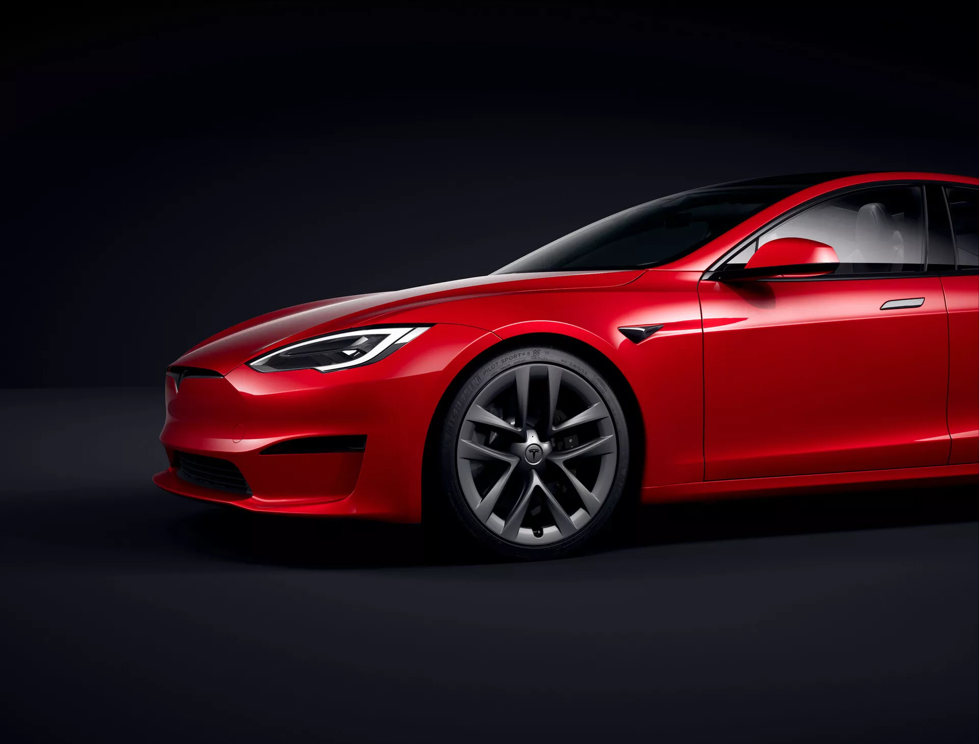 Tesla Model S 2016 (prenova prve generacije) rdeča limuzina v studiu z nadzorovano osvetlitvijo in črnim ozadjem, nizek kotni pogled, ki poudarja aerodinamični profil in oddaljenost od tal, tridelni pogled od spredaj, ki prikazuje stran voznika, žaromete, rešetko, del pokrova motorja, platišče iz premium zlata z večkrakim vzorcem, vetrobransko steklo s toniranim steklom, nagnjena linija strehe, stransko ogledalo, kljuke vrat in linija okna, ki se gladko vključujejo v eleganten dizajn, poudarjajoč zaključek barve, konturne črte in oblikovane plošče, ki nakazujejo luksuz in zmogljivost, osredotočena osvetlitev, ki prikazuje dizajnerske podrobnosti.