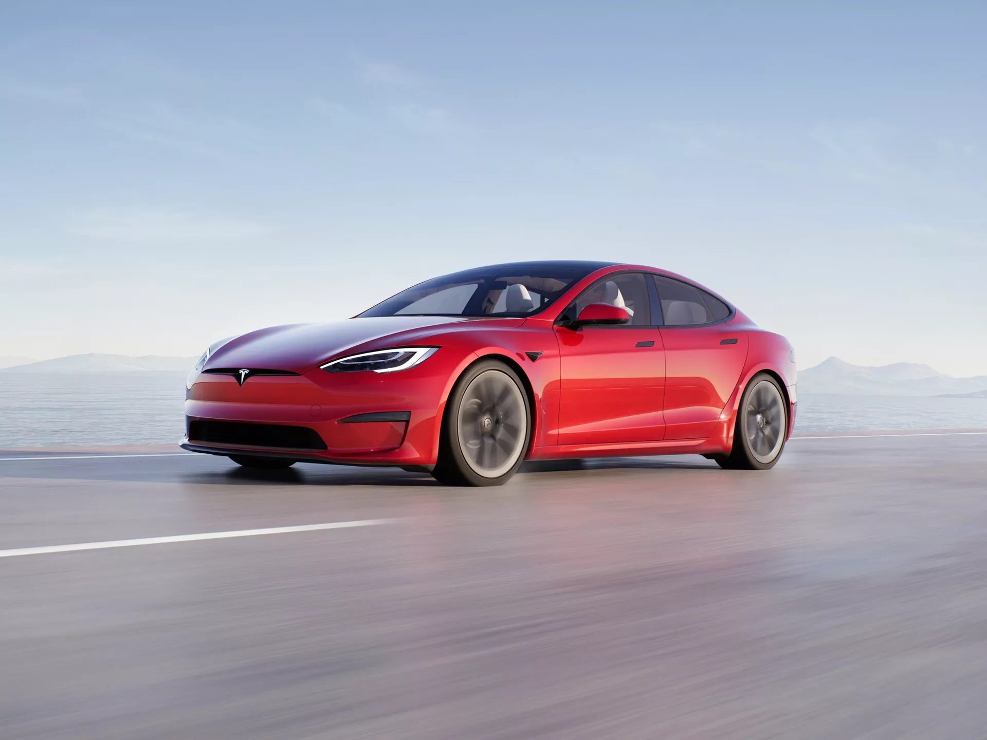 Slika prikazuje sodoben štirivratni limuzinski avto Tesla Model S 2016 (1. generacija facelift) v gibanju na gladki cesti ali stezi. Vozilo je predstavljeno iz nizkega kota stranskega profila, tako da so jasno vidne elegantne in aerodinamične linije, vključno z značilno zaprto masko, tanjimi LED žarometi in gladkim sprednjim odbijačem. Fastback dizajn z nežno padajočo linijo strehe, vgrajenimi uvlačljivimi kljukami vrat in minimalističnimi aerodinamičnimi stranskimi ogledali poudarja osredotočenost vozila na aerodinamiko. Velika večkraka kolesa nakazujejo optimizacijo za pretok zraka in zmanjšanje zračnega upora. Kompozicija uporablja zameglitev gibanja, da bi prikazala visoko hitrost, pri čemer je avto sam ostro izostren proti zamegljenemu ozadju. Ta profesionalna fotografija poudarja električno naravo avtomobila, napredno osvetlitev, aerodinamičen dizajn in potencialne funkcije pomoči vozniku.