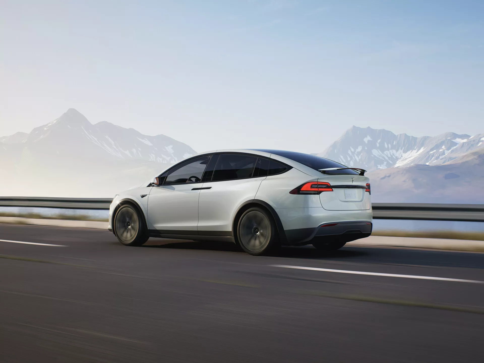 Dinamičen zadnji tričetrtinski pogled na vozilu Tesla Model X 2021 (1. generacija-facelift) v gibanju, ki poudarja njegovo aerodinamično zasnovo, elegantno belo barvo, LED zadnje luči in sodobno obliko koles. Vozilo je ujeto med premikanjem od leve proti desni na zamegljeni cesti in ob ograji, kar nakazuje hitrost, v ozadju pa je gorski obzorje. Mehka osvetlitev kaže na zgodnje jutro ali pozno popoldne, kar poudarja čiste električne linije in napredne funkcije vozila v slikovitem okolju.
