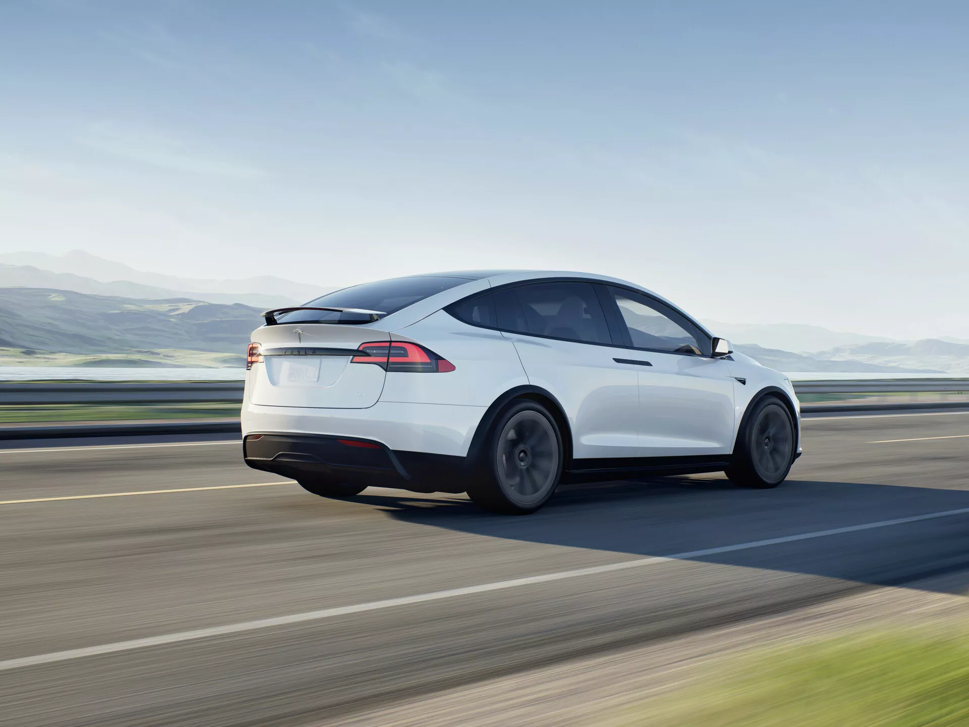 Dinamična slika belega električnega hatchback sedana Tesla Model X 2021 v gibanju, prikazana iz zadnjega tričetrtinskega kota. Fotografija ujame levi bok vozila iz rahlo dvignjenega kota, poudarja zadnja kolesa, zadnje luči in obris zadnjega dela avtomobila, poudarja njegovo aerodinamično obliko z nagnjeno linijo strehe in gladkimi krivuljami. Oblikovanje nakazuje zmanjšano zračno turbulenco in nizek količnik upora, podprto s kolesi, oblikovanimi za aerodinamično učinkovitost. Zadnje okno se zlije v črno stekleno streho, kar nakazuje prisotnost panoramske strešne okna, simbol moderne avtomobilske tehnologije. Elegantne LED zadnje luči so značilnost sodobne svetlobne tehnologije. Odsotnost izpušne cevi kaže na popolnoma električno naravo vozila. Idealno za kategorije, kot so vozila, električna vozila, fotografija gibanja, avtomobilsko oblikovanje, moderna tehnologija in luksuzna vozila.