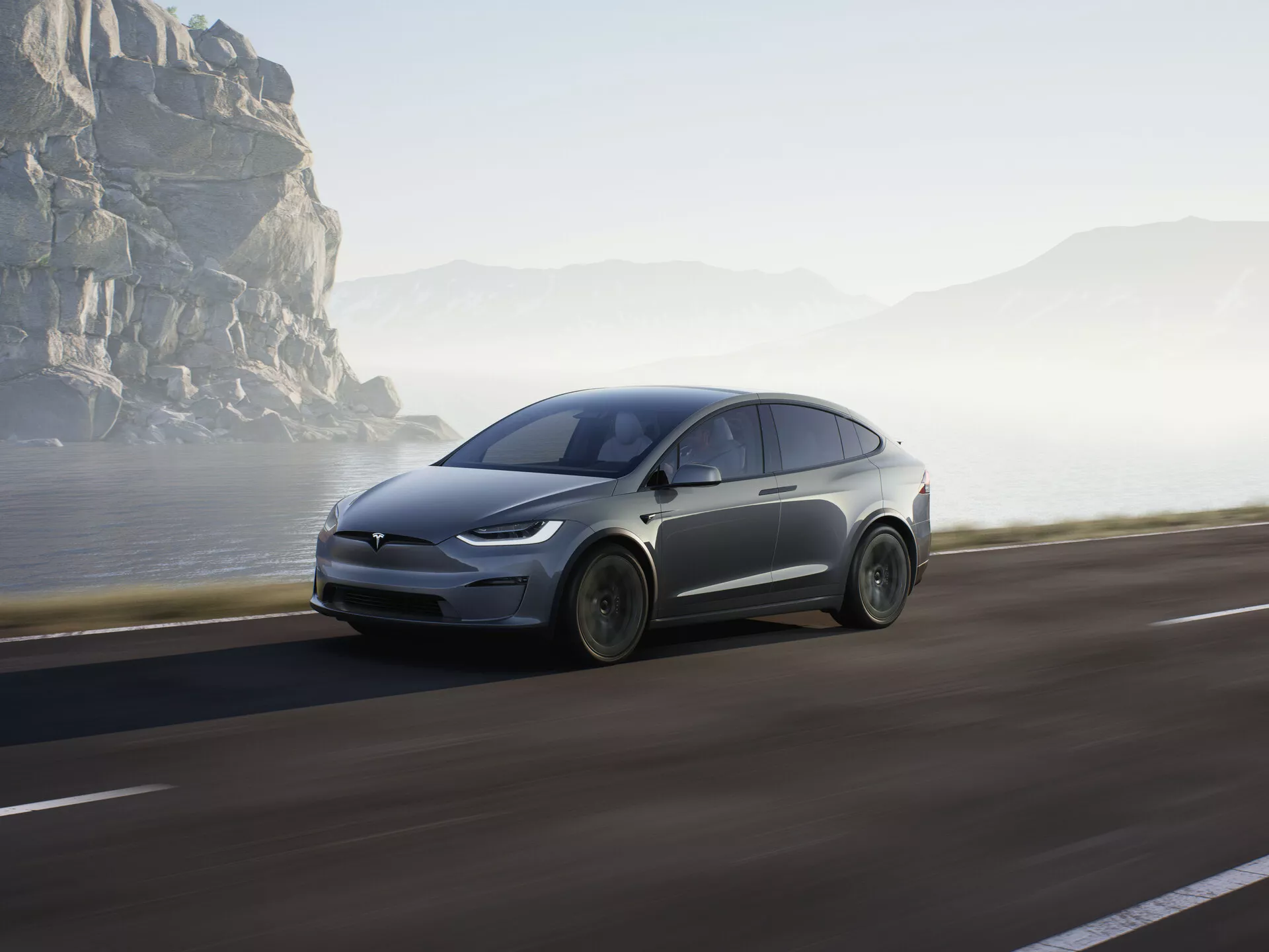 Slika prikazuje sodoben električni SUV, natančneje Tesla Model X 2021 (1. generacija facelift), ki vozi po obalni cesti. Posnet je iz nizkega stranskega profila, ki poudarja sprednjo četrtino s poudarkom na sprednjem kolesu, pokrovu motorja, vetrobranskem steklu in delu voznikovih vrat. Vozilo zaseda pomemben del spodnje polovice okvirja, kar poudarja njegovo izrazitost. Avto je v gibanju, kar dokazuje rahla zameglitev koles in ozadja, ustvarjajoč občutek hitrosti in dinamike. Mehka sončna svetloba nakazuje zgodnje jutro ali pozno popoldne, kar prizoru doda umirjeno vzdušje v slikovitem in pustolovskem obalnem okolju. Predvidevati je mogoče, da so prisotni aerodinamični oblikovalski elementi in napredne funkcije, kot so LED žarometi, kar nakazuje napredno tehnologijo, kot so regenerativno zaviranje in morda avtonomne vožnje zmogljivosti. Avto je nameščen na desni strani ceste in se premika naprej proti odprtemu cilju, pri čemer obalne skale in megla v ozadju poudarjajo njegov futurističen videz in naravno okolje.