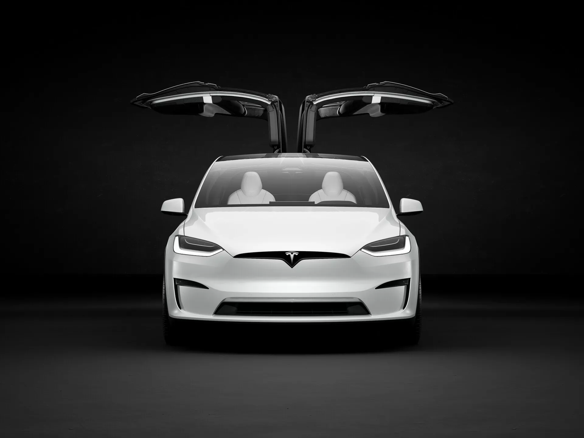 Slika prikazuje električno vozilo Tesla Model X 2021 (prva generacija, prenova) v studijskem okolju s temnim ozadjem, ki kontrastira s svetlo barvo avtomobila. Pogled je rahlo iz nizkega kota, izpostavljajoč sprednji del avta, vključno s pokrovom motorja, odbijačem in režo za prezračevanje, čeprav ta ni tradicionalna, ker je električno vozilo. Vidna je tudi spodnja reža za zrak. Oboja sprednja vrata sta odprta, razkrivajoč edinstvena vrata falcon-wing, ki se dvignejo navzgor, kar je značilnost visoko kakovostnih modelov. Notranjost prikazuje dve vrsti svetlih sedežev, kar nakazuje minimalistični, a luksuzni dizajn. Ostre, kotne LED luči dodajajo agresiven videz, logotip Tesla pa je vidno nameščen nad območjem za zajem zraka in pod 'frunkom'. Avtomobil namiguje na napredne funkcije, kot so avtonomna vožnja, električni pogonski sistem, regenerativni zavorni sistemi, povezane lastnosti avtomobilov in napredni varnostni sistemi, namenjeni občinstvu, ki ceni inovacije, zmogljivost in okoljsko zavest.