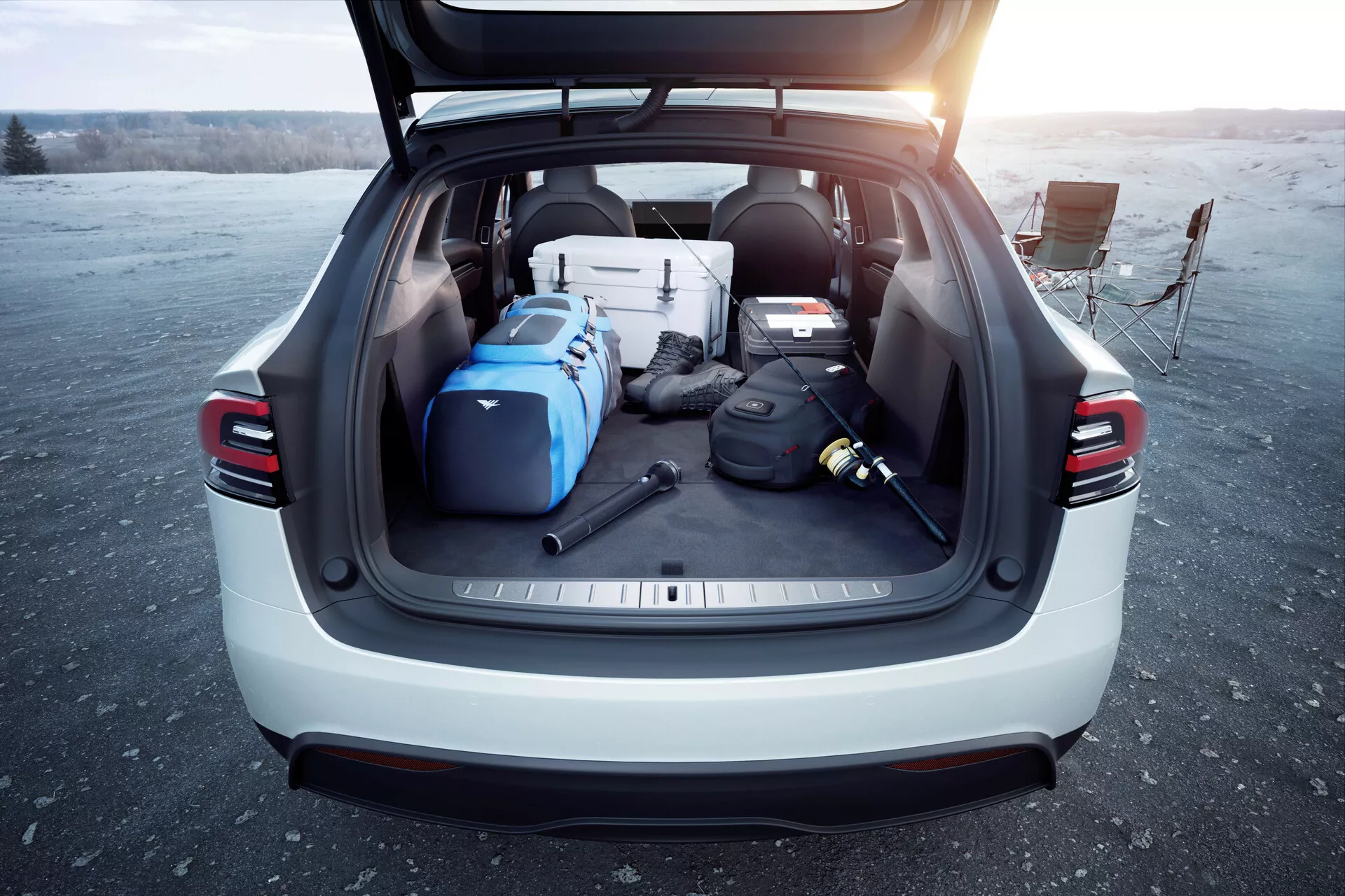 Pogled s hrbtne strani SUV Tesla Model X 2021 z odprtim prtljažnikom, ki prikazuje prostoren tovorni prostor, napolnjen z različnimi predmeti pod dnevno svetlobo, morda ob sončnem vzhodu ali zahodu. Slika poudarja uporabnost in sodobne lastnosti vozila, kot so avtomatizirana prtljažna vrata, zadnje LED luči, svetla zunanja barva in pokrov za tovor, posneta na odprtem prostoru.