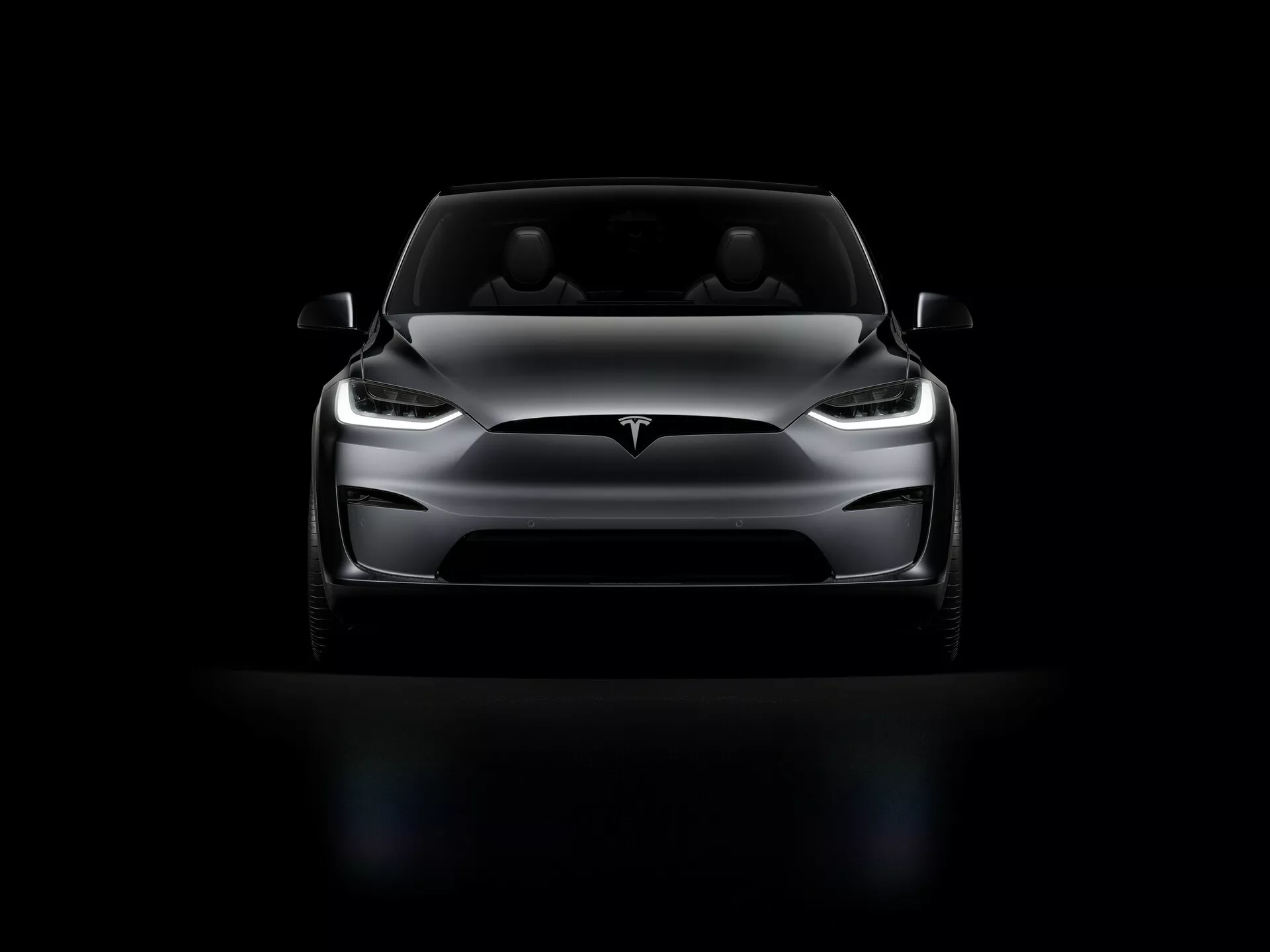 Tesla Model X 2021 (prva generacija facelift) fotografirana iz sprednjega kota pod nizkim kotom. Slika poudarja sprednji del avtomobila, LED žaromete, eleganten dizajn pokrova motorja, gladek sprednji del brez mreže, minimalistični sprednji odbijač in aerodinamična bočna ogledala. Dramatična osvetlitev poudari ključne značilnosti, medtem ko avto stoji na črnem ozadju, kar povečuje njegov impozantni videz.