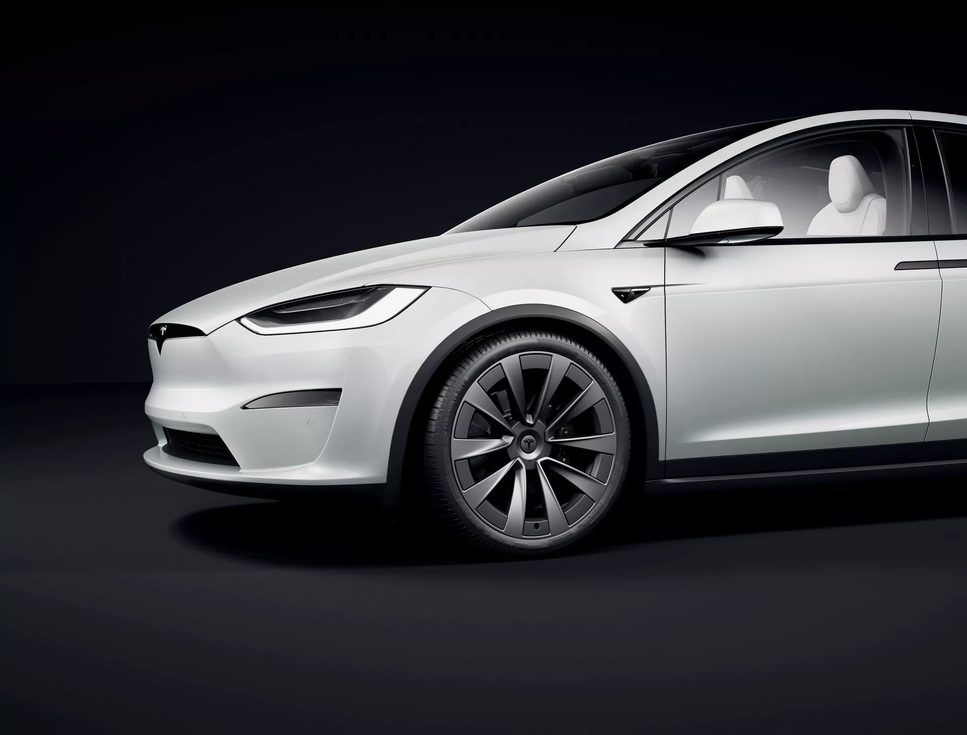 Studijska slika, ki prikazuje stranski profil prenovljenega modela Tesla Model X 2021 prve generacije iz nizkega kota, ki poudarja luksuzne električne funkcije, kot so elegantna zasnova, odsotnost izpušnih cevi, minimalistična sprednja maska z LED žarometi, uvlečne kljuke vrat, večkraka aero kolesa, velike nizkoprofilne pnevmatike, sijajna bela barva z mehkimi odsevi, panoramska streha in kamere namesto stranskih ogledal. Krivulje in linije vozila poudarja studijska osvetlitev proti temnemu gradientnem ozadju.