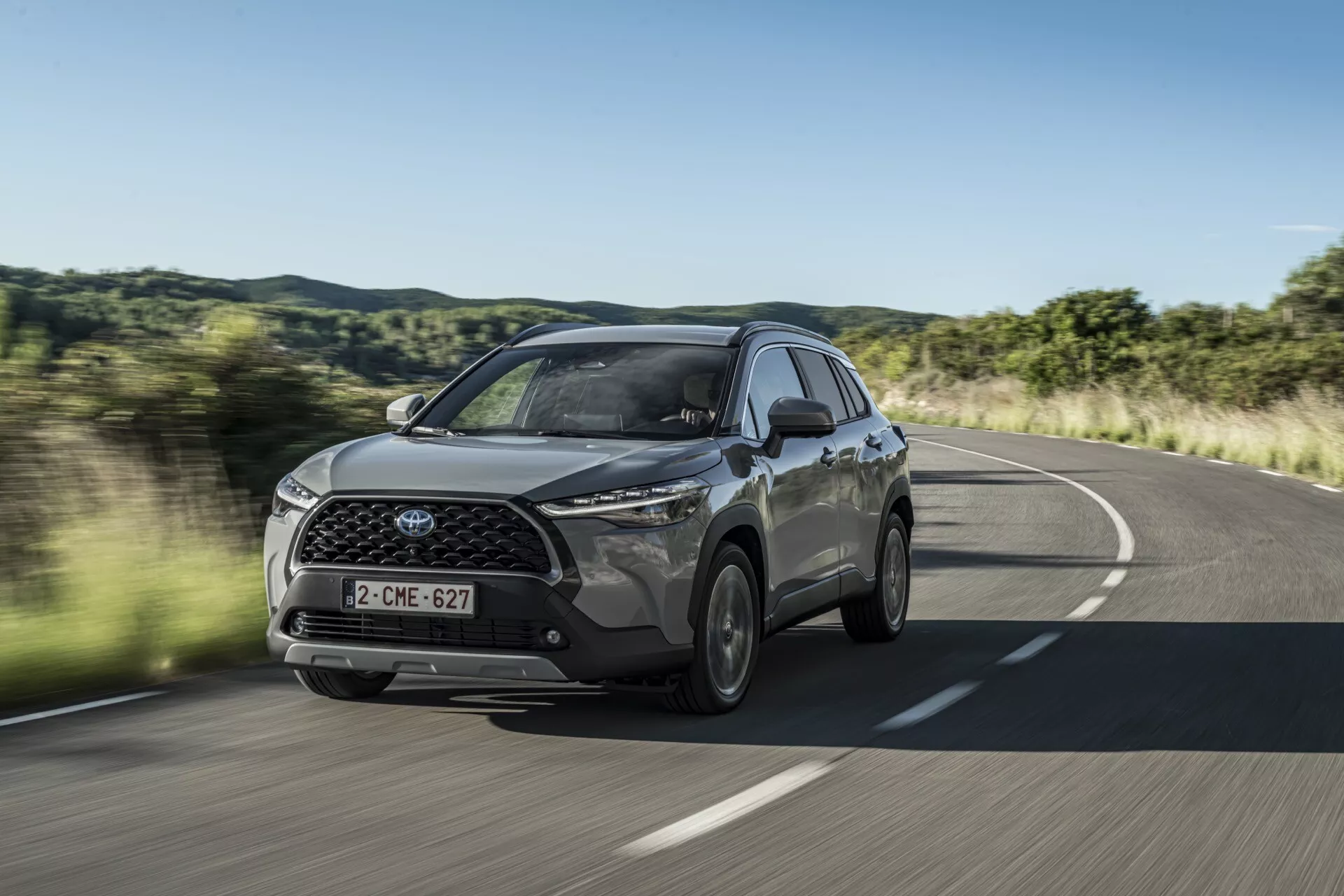 Model Toyota Corolla Cross prve generacije leta 2022 v gibanju na podeželski cesti s pogledom spredaj in s strani, ki poudarja LED žaromete, sprednjo rešetko z emblemo in stranske lita kolesa