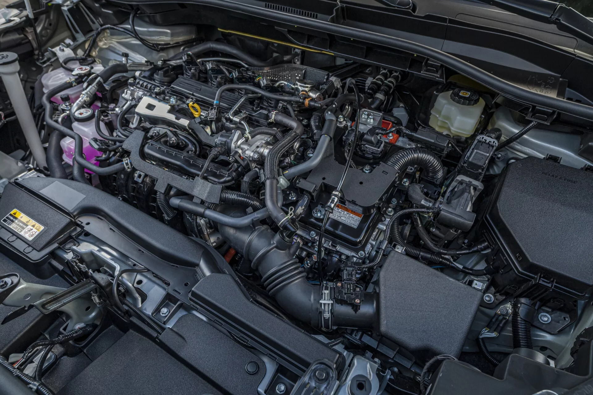 Podroben pogled na motorni prostor Toyota Corolla Cross 2022 (1. generacija), ki poudarja njen sodobni motor z notranjim zgorevanjem in različne avtomobilske tehnologije, kot so elektronski sistem upravljanja motorja, pokrov motorja za zatiranje hrupa, rezervoarji za tekočine, sistem za dovod zraka in strateške komponente za upravljanje toplote.