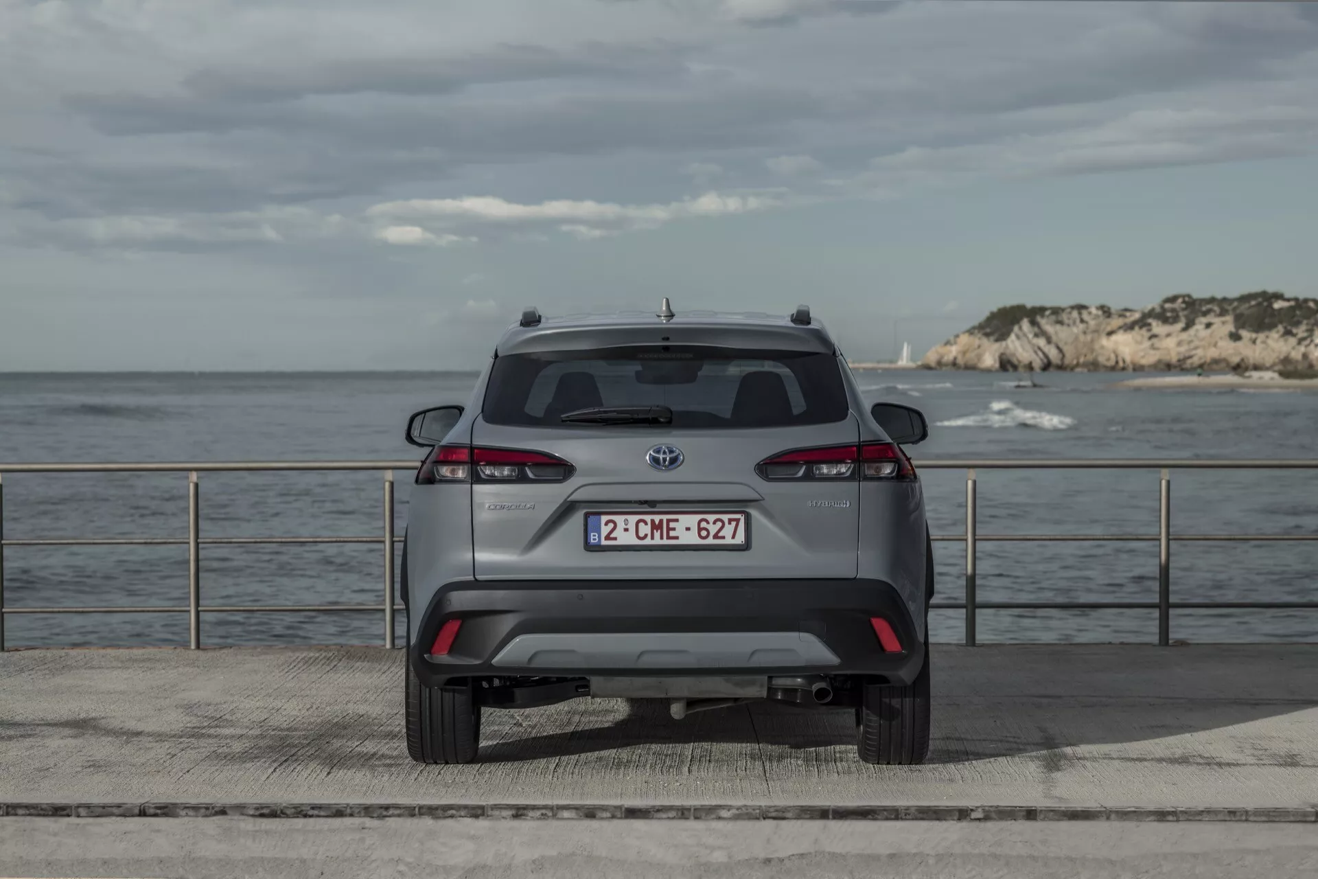 Zadnji pogled na Toyota Corolla Cross 2022, parkirana na obalni platformi z naravno osvetlitvijo, ozadjem obzorja, popolnimi zadnjimi lučmi, zadnjim vetrobranskim steklom, brisalcem, znamko na zadnjem odbijaču, modelno značko, registrsko tablico in tehnološkimi varnostnimi elementi, kot so senzorji za parkiranje.