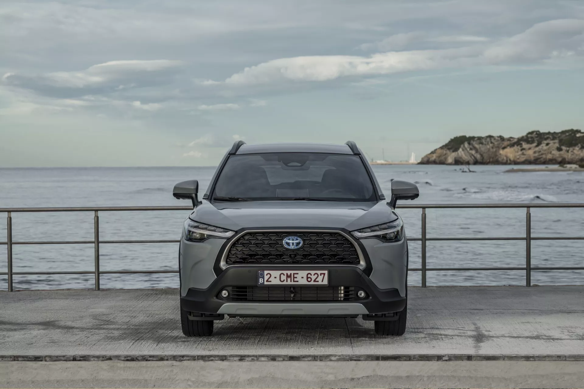 Sprednji pogled na Toyota Corolla Cross 2022, ki prikazuje simetrično zasnovo, napredne LED žaromete, zapleteno rešetko z emblemo Toyota in sodobne zunanje značilnosti proti nevtralnem ozadju z vodo in oblačnim nebom