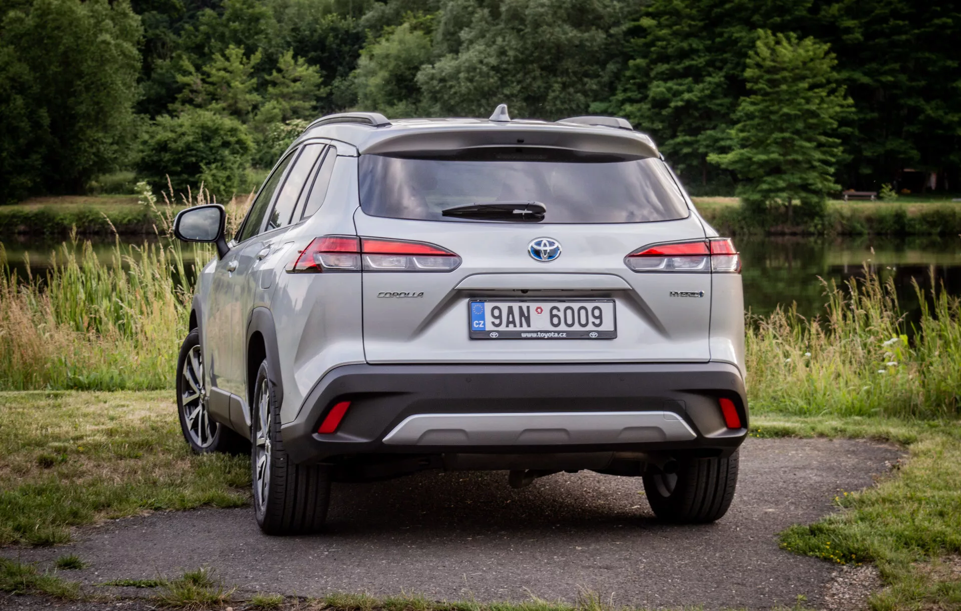 Srebrni Toyota Corolla Cross letnik 2022, parkiran na tlakovcu z zelenjem v ozadju, prikazuje svoje LED zadnje luči, zadnji spojler, večkrako zlitinsko kolo in hibridno oznako ob dnevni svetlobi