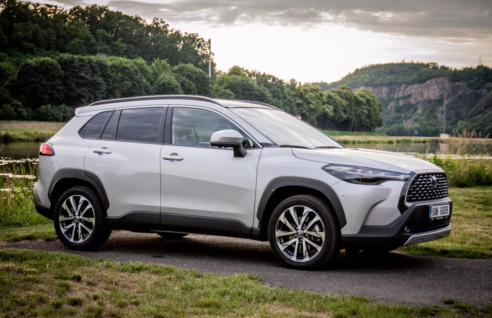 SUV Toyota Corolla Cross 2022, parkiran na prostem, ki s treh četrtinskimi čelnimi koti prikazuje svoj sodobni dizajn s poudarkom na mreži hladilnika, platiščih iz lahke litine in stranskem profilu proti naravnemu ozadju