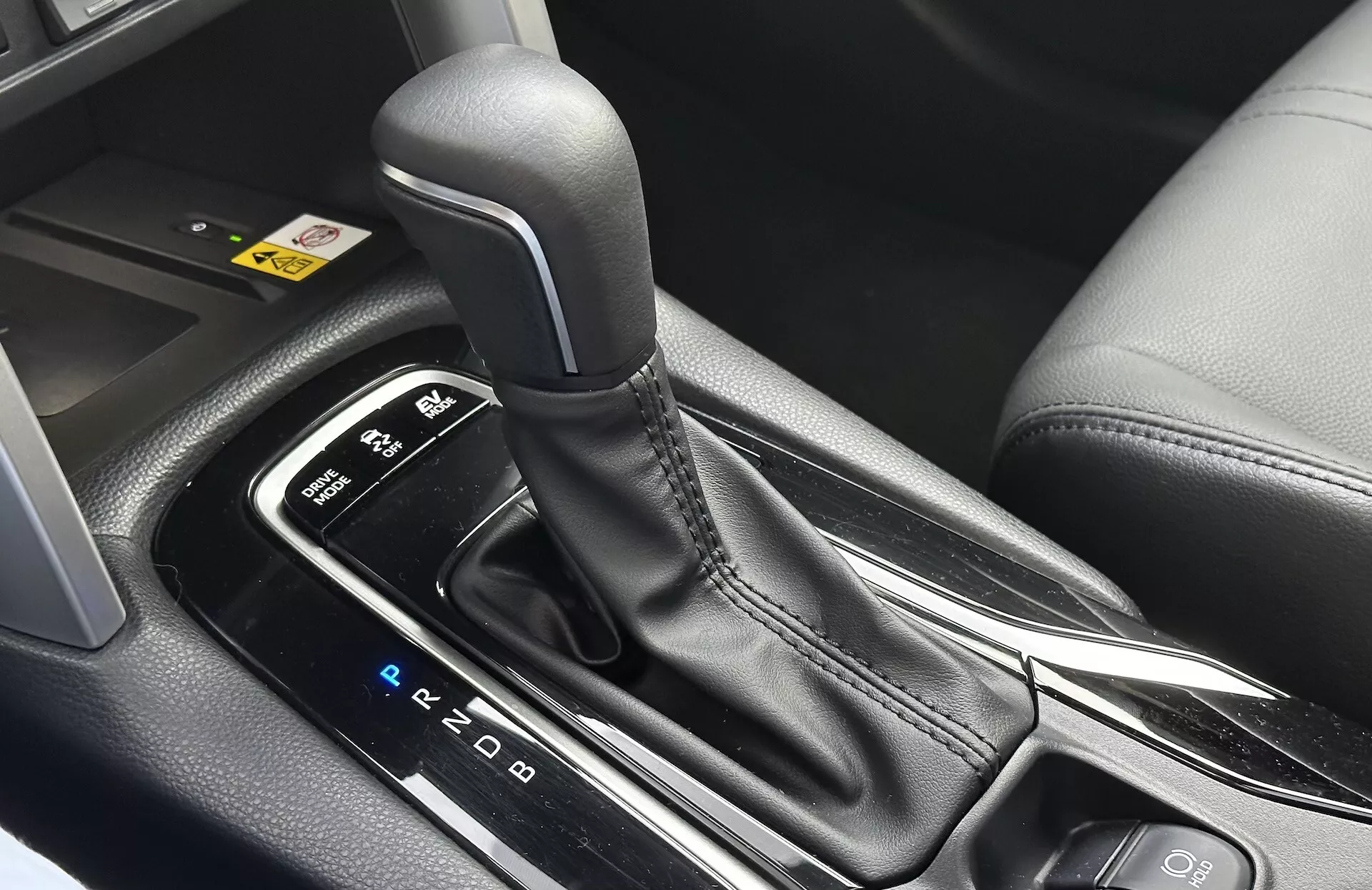 Podroben notranji pogled na Toyota Corolla Cross 2022, ki prikazuje avtomatsko prestavno ročico s vidno postavitvijo PRND, obdano z elegantno sredinsko konzolo z črnimi plastičnimi in kovinskimi poudarki, poudarjajoč ergonomski dizajn in sodobne tehnološke značilnosti