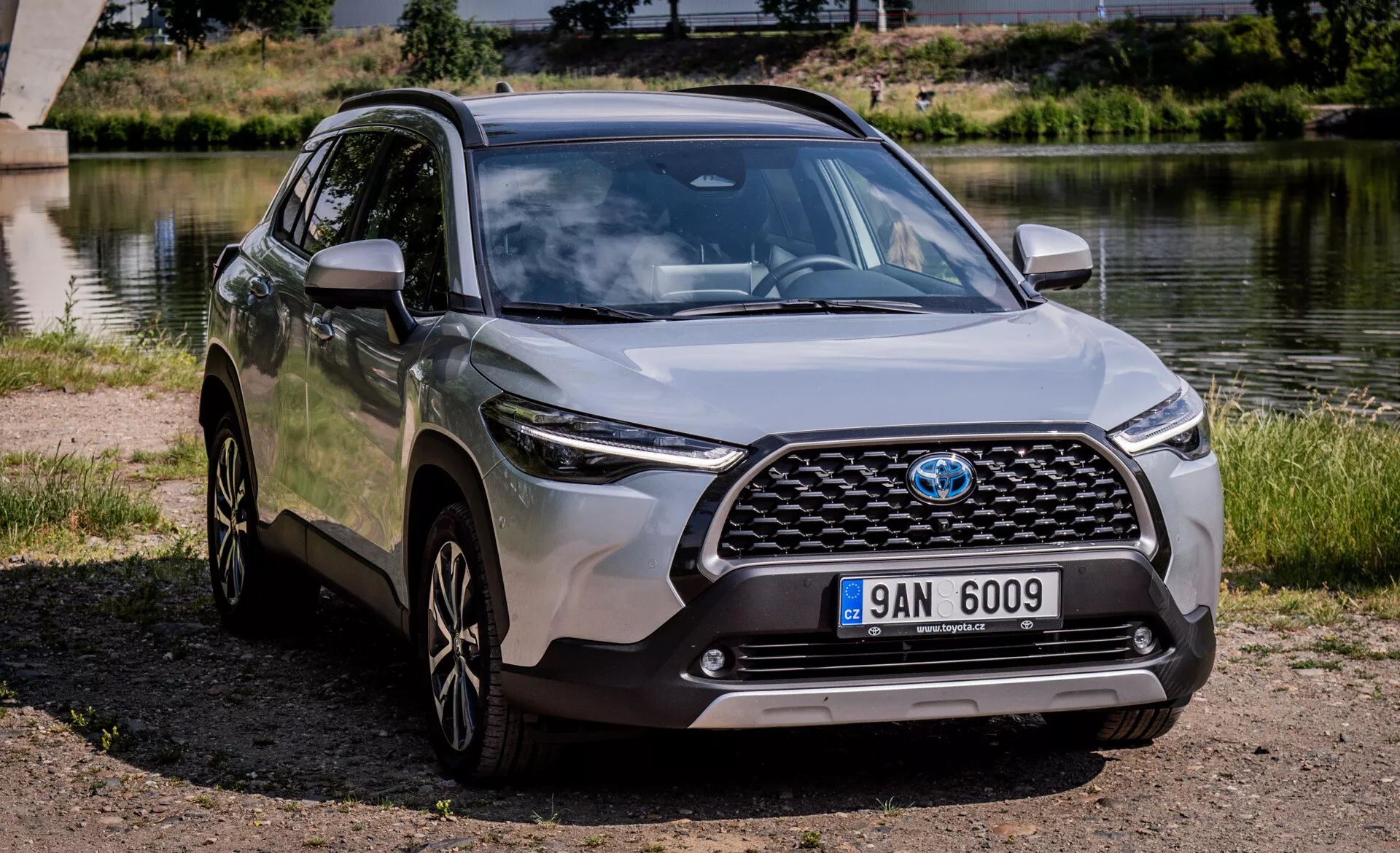 Sodoben crossover SUV Toyota Corolla Cross 2022 parkiran na prostem blizu vode z jasnim pogledom na njegovo sprednjo masko, pokrov motorja, LED žaromete, sprednji odbijač in stranski profil vključno z litimi platišči in bočnimi ogledali v barvi karoserije