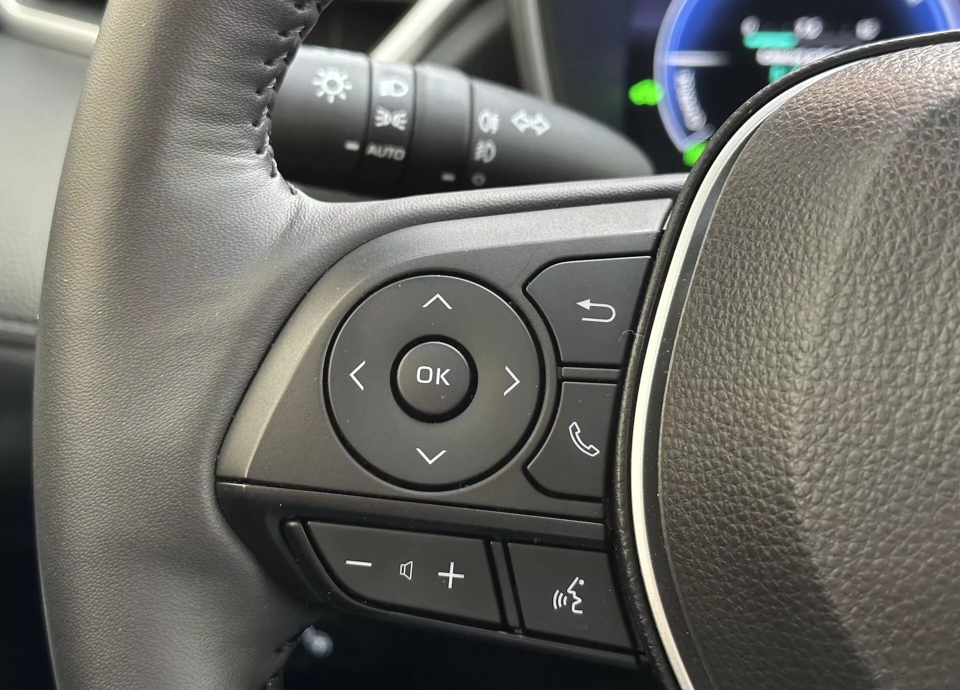 Podroben pogled na volan Toyote Corolla Cross 2022, osredotočen na večnamenske kontrole na levem kraku z usnju podobno teksturo in jasno označenimi gumbi za avdio, Bluetooth telefonsko povezavo, glasovne ukaze in navigacijo preko sistema informacij voznika