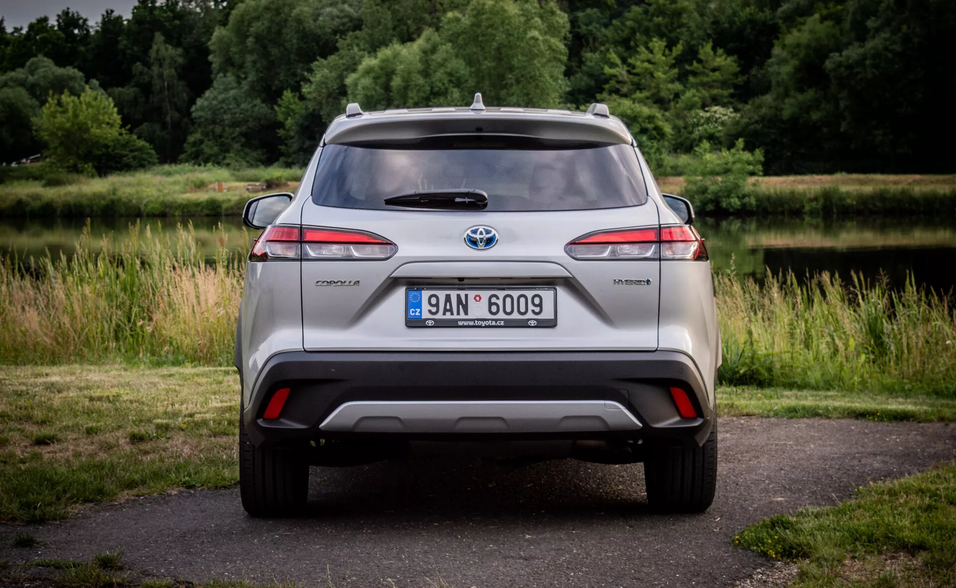 Zadnji pogled na hibridni model Toyota C-HR z LED zadnjimi lučmi, integriranim odbijačem z difuzorjem in odsevniki, anteno nameščeno na strehi in litoželeznimi kolesi, postavljen proti naravnemu ozadju