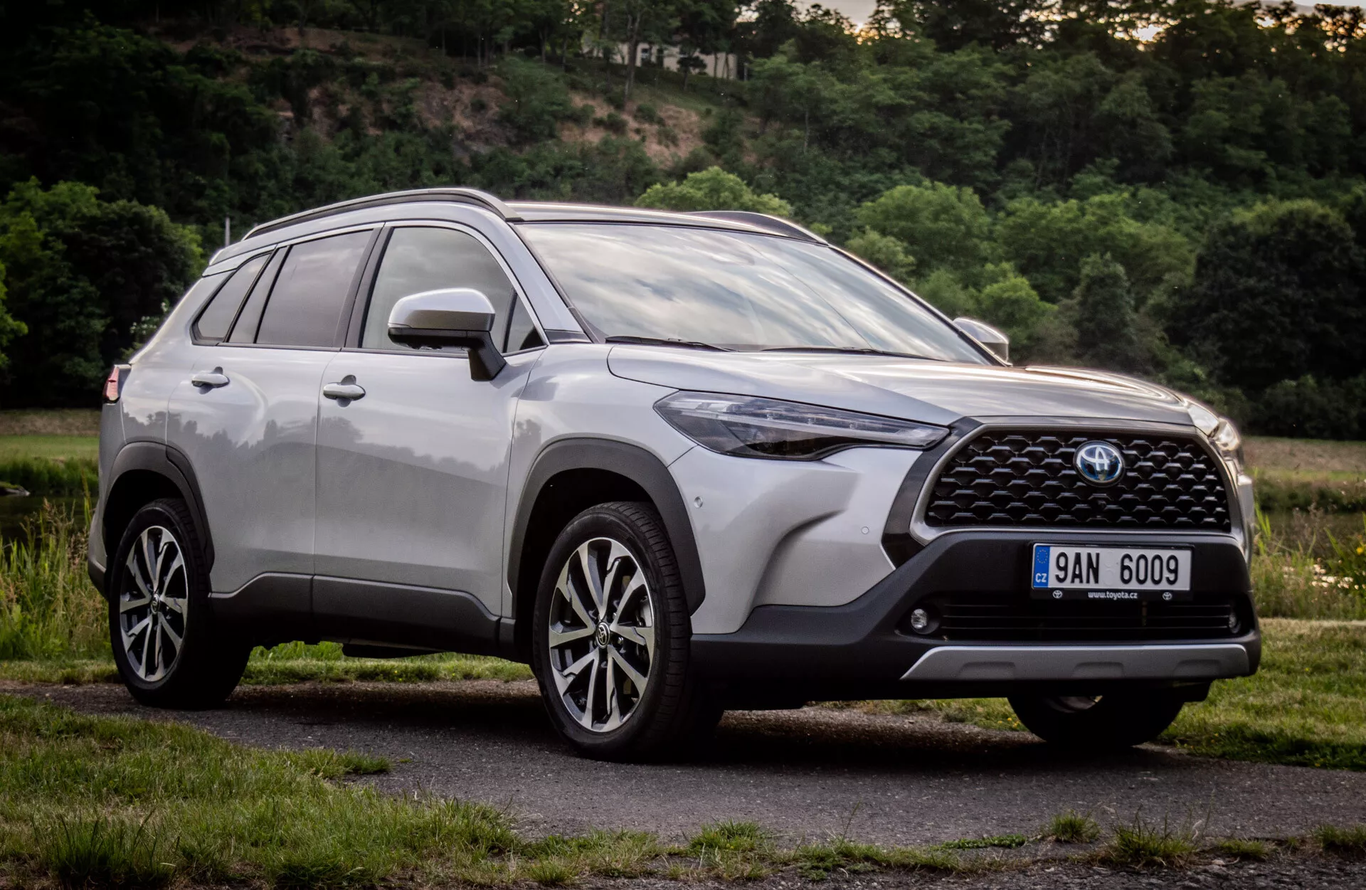 Toyota Corolla Cross 2022 v sijočem kovinskem zaključku, ki prikazuje sodobno zasnovo SUV z natančno sprednjo masko, emblemom, kotnimi LED žarometi, aerodinamičnim sprednjim odbijačem, konturiranim pokrovom motorja, stranskim profilom z večkrakimi litimi kolesi in strešnimi nosilci, posnetim iz nizkega kota, da poudari njeno višino in držo podnevi v naravnem predmestnem okolju.