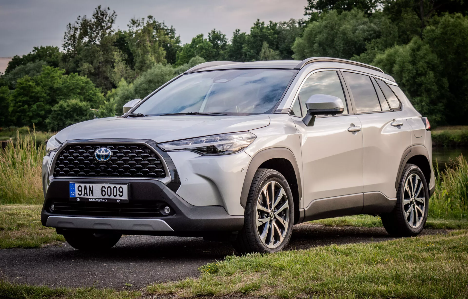 Toyota Corolla Cross 2022 prikazuje svojo dinamično obliko s pogledom spredaj in po strani, poudarja veliko vzorčasto sprednjo rešetko, vitke LED žaromete in večkraka lita kolesa, parkirana v naravnem parku
