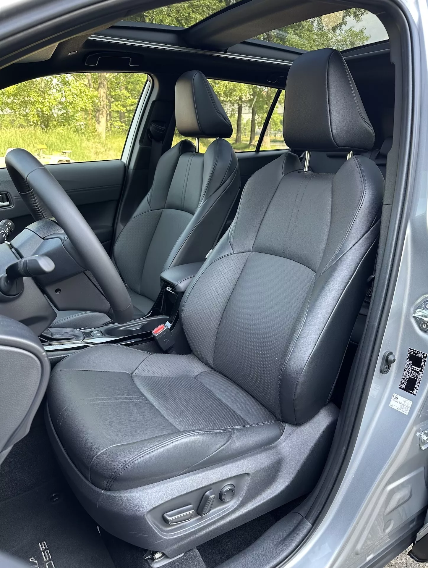 Notranjost Toyota Corolla Cross 2022 z izpostavljenimi črnimi usnjenimi sprednjimi sedeži z nastavitvenimi kontrolami, delom zadnjih sedežev, volanom in varnostnimi značilnostmi okvirja vrat v naravni dnevni svetlobi