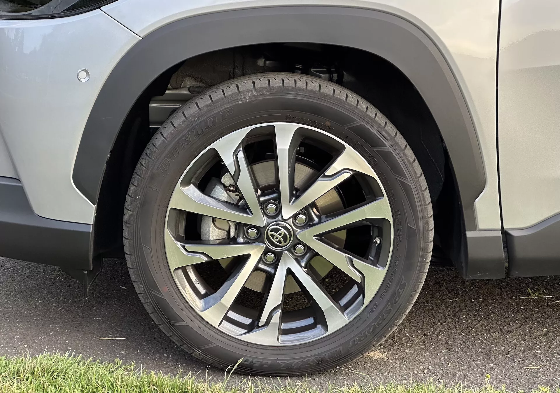 Podroben posnetek desnega sprednjega kolesa Toyota Corolla Cross 2022 z večkrakim dizajnom iz lahke zlitine, profilom pnevmatike za vse letne čase in brezhibno karoserijo SUV-ja, parkiranim na asfaltu.