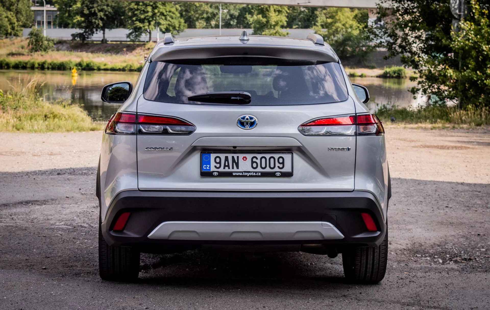 Zadnji pogled na Toyota Corolla Cross 2022, prikazuje LED zadnje luči, hibridno značko in kromirani emblemi Toyota s naravno dnevno svetlobo in slikovitim ozadjem zelenja in vode