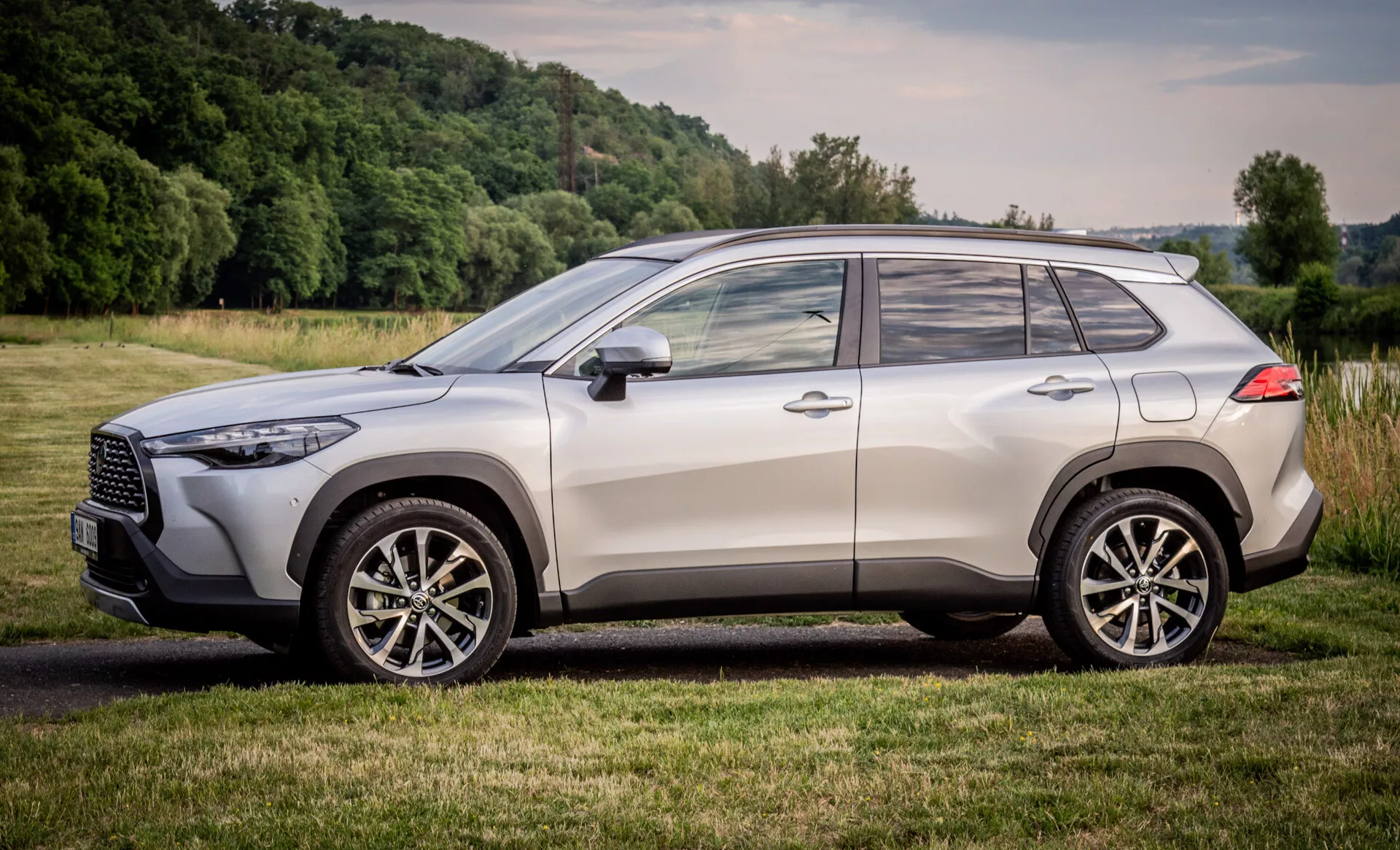 Toyota Corolla Cross 2022, parkirana na travnatem prostoru, z bočnim profilom, sodobno oblikovanje SUV z okraski iz kroma, platišči iz zlitin in napredno tehnologijo osvetlitve