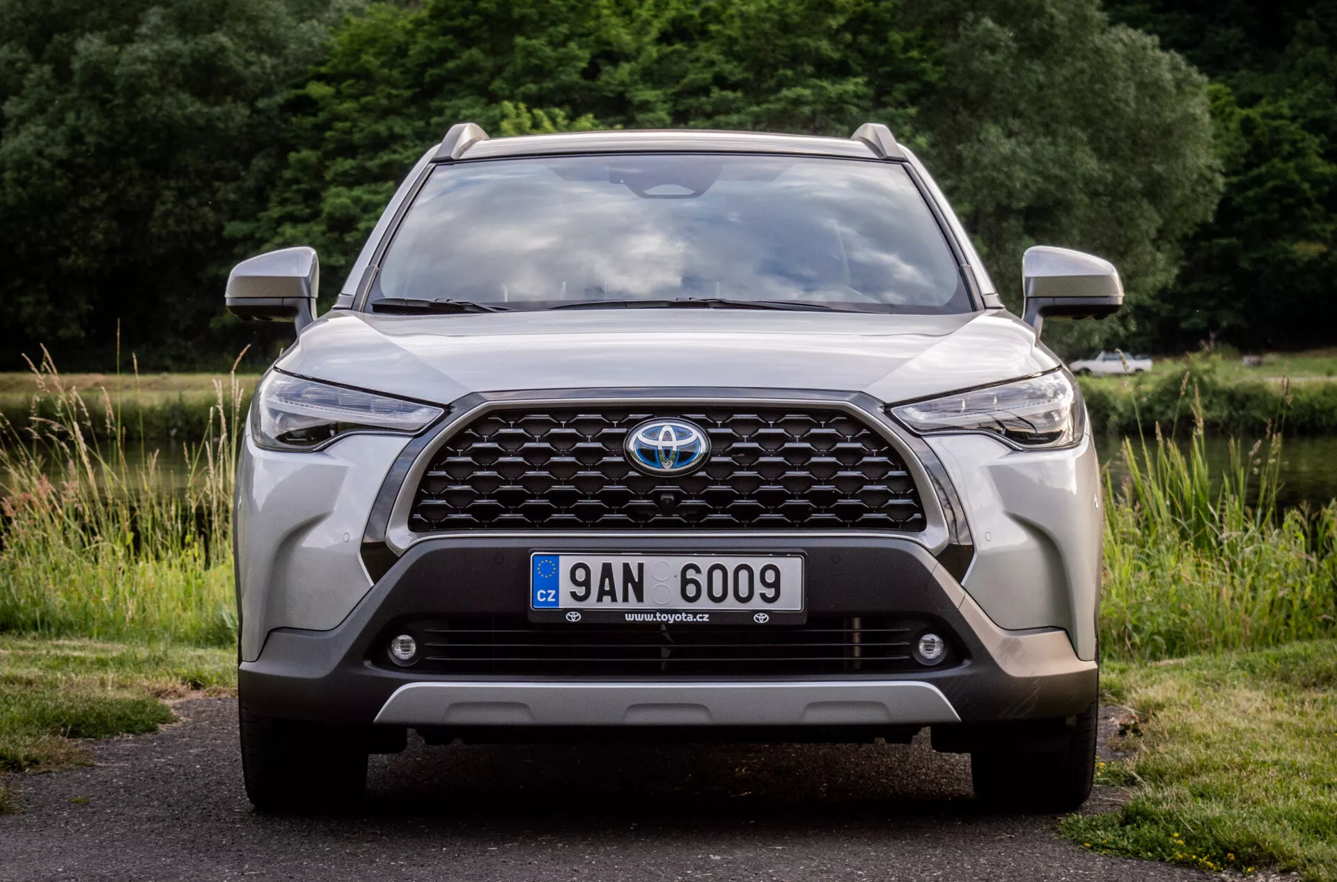 Sprednji pogled na Toyota Corolla Cross 2022, ki poudarja njeno veliko rešetko z emblemo, sodobne LED žaromete, meglenke, pozicijo registrske tablice, integrirane senzorje za parkiranje ali sprednjo kamero, aerodinamične konture pokrova motorja, robustno oblikovanje spodnjega odbijača in zračne vhode, parkirano na asfaltu z naravnim ozadjem