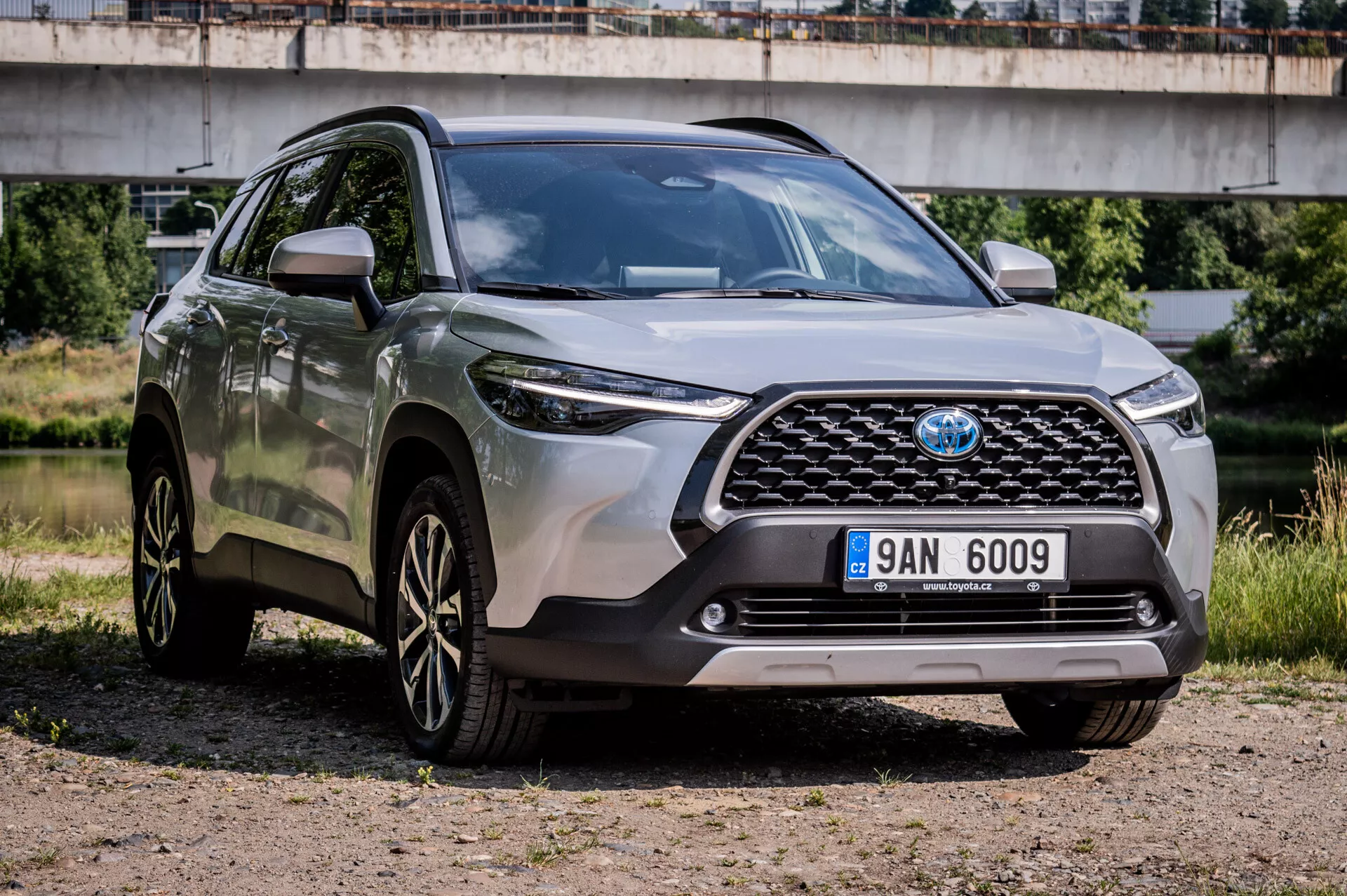 Toyota Corolla Cross 2022, parkirana na prodnatem terenu, prikazuje svoj robustni dizajn, športna lita platišča z več žarki, napredne LED žaromete in visoko oddaljenost od tal v pogledu spredaj treh četrtin, ki poudarja sprednjo rešetko, odbijač in lastnosti na voznikovi strani z mostom v polurbani ozadju