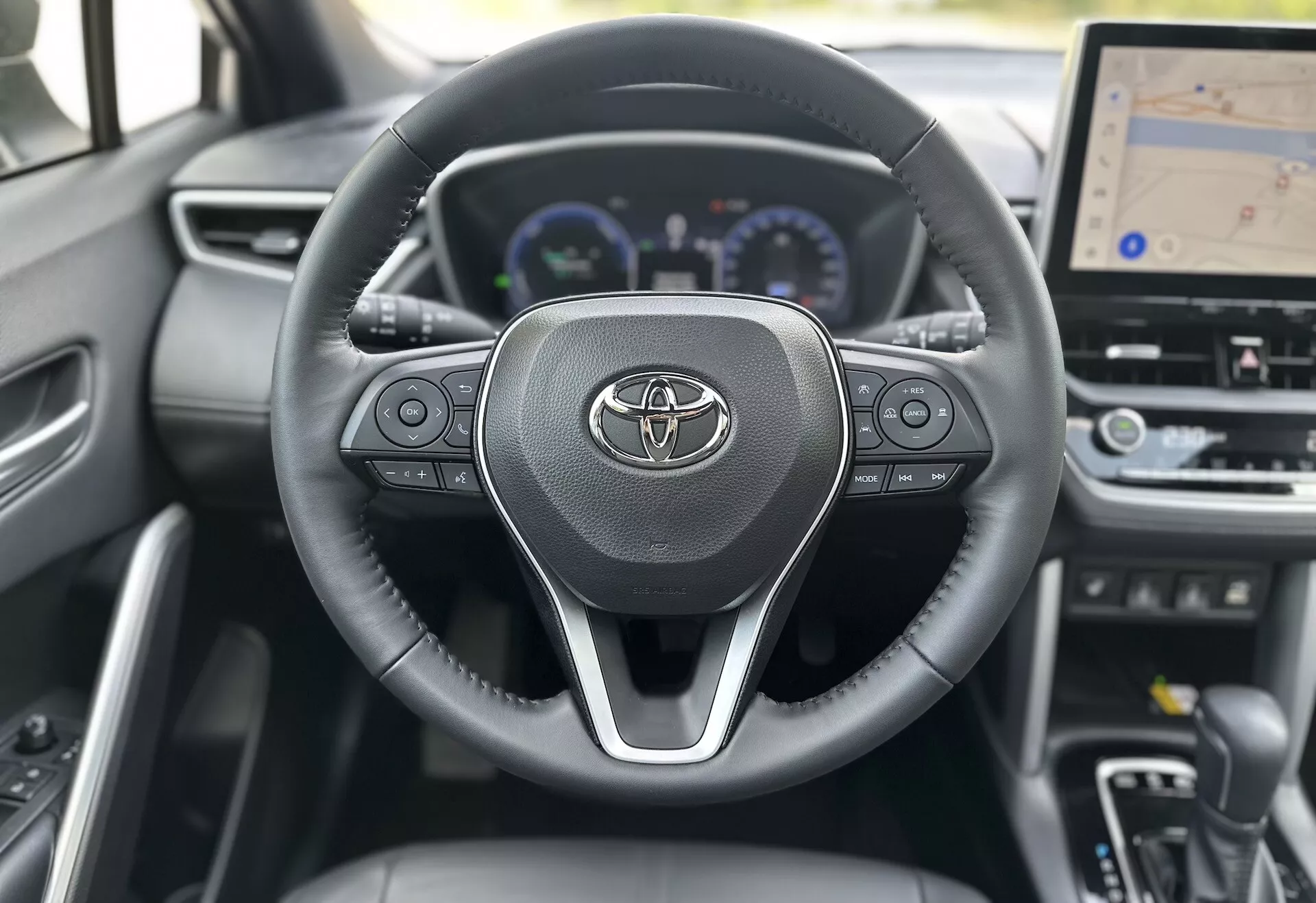 Notranjost Toyote Corolla Cross 2022, ki prikazuje volan z logotipom Toyote, vgrajenimi kontrolami za avdio, klice in tempomat, za katerim je delno viden digitalni armaturna plošča z merilnikom hitrosti, tahometrom in večfunkcijskim zaslonom, vključno z večjim osrednjim zaslonom armaturne plošče za GPS navigacijo, vse na ozadju sodobnega dizajna iz temnih materialov.