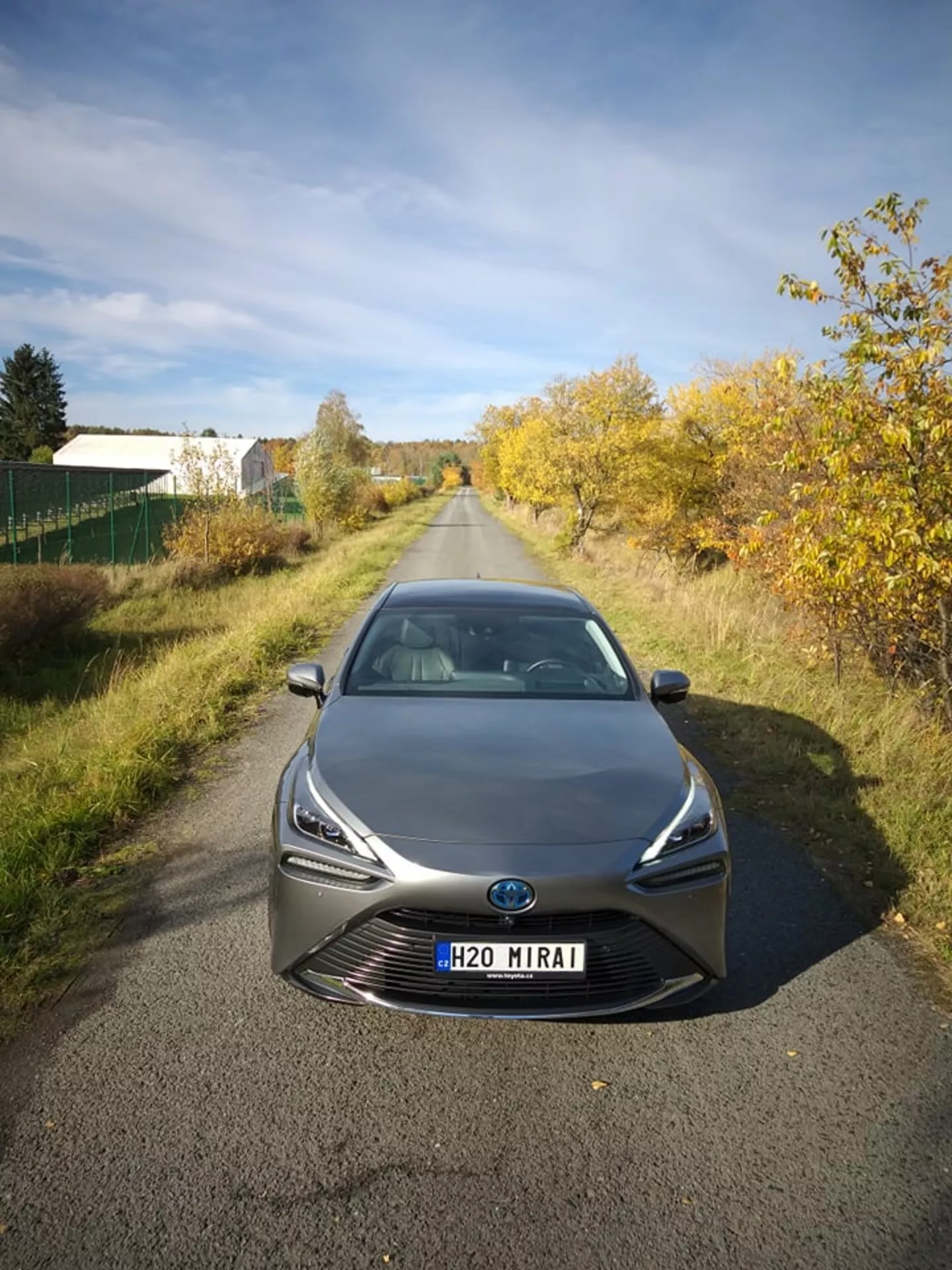 Toyota Mirai 2021, fotografirana s sprednje strani pod rahlo povišanim kotom, ki prikazuje njene elegantne LED žaromete in izpostavljeno masko z osrednjim emblemom, kar morda vključuje senzorsko tehnologijo, v naravni dnevni svetlobi na mirni cesti z drevesi, kar nakazuje, da je vozilo na predmestnem ali polzemeljskem območju, predstavljajoč napredno tehnologijo gorivnih celic na vodik in aerodinamično oblikovanje