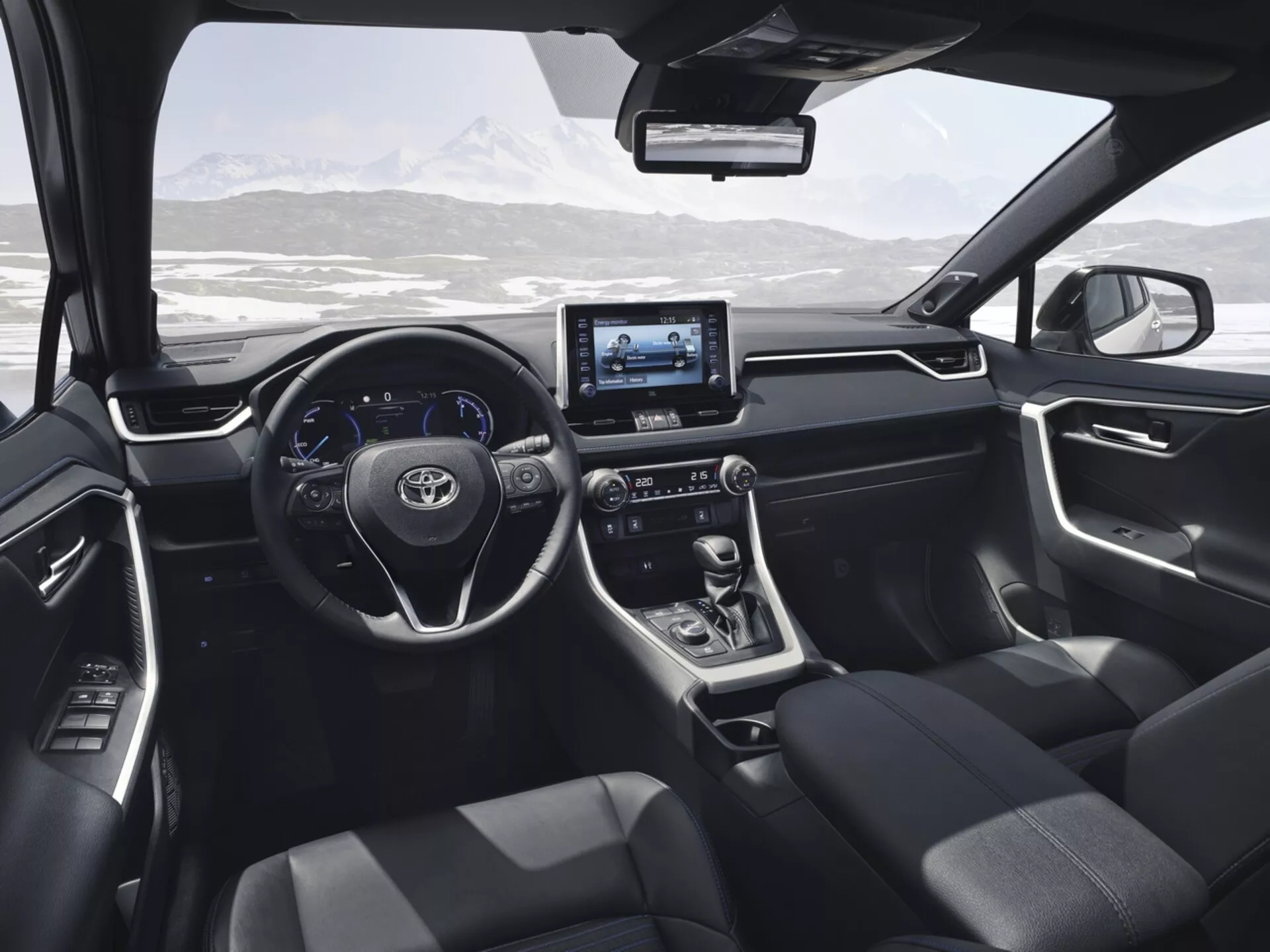 Pogled na notranjost Toyota RAV4 2019 iz zadnjega sedeža, ki predstavlja sodobno oblikovanje in tehnološke funkcije, vključno z volanom z nadzornimi gumbi, digitalnim zaslonom, zaslonom na dotik za informacijsko-zabavni sistem, ročico za avtomatski menjalnik, temno oblazinjenimi sedeži in nadzornimi ploščami na vratih z razgledom na snežno pokrajino z gorami skozi vetrobransko steklo.