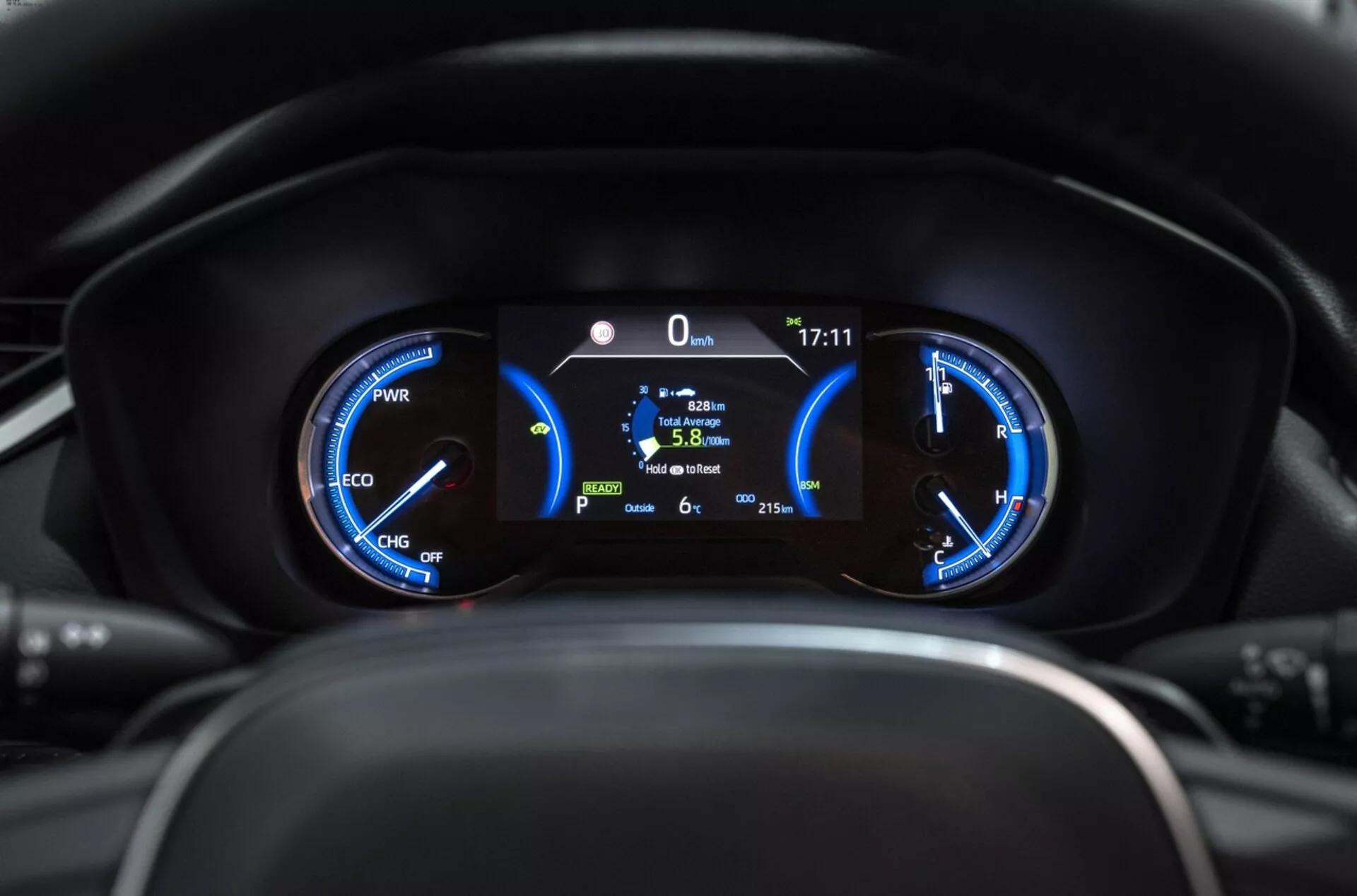 Posnetek merilnikov Toyota RAV4 2019 iz bližine, prikazuje hibridne merilnike z merilnikom moči, hitrostomerom in centralnim digitalnim zaslonom, ki prikazuje stacionarno pozicijo, učinkovitost porabe goriva in zunanjo temperaturo iz perspektive voznika.