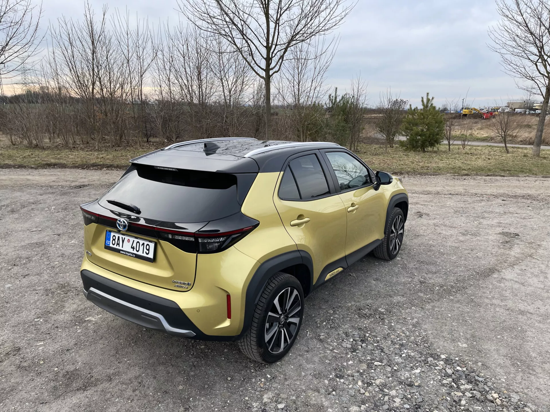 Toyota Yaris Cross 2021 ujeta s treh četrtinskega zadnjega pogleda, ki poudarja njen aerodinamični spojler, metalik zlato barvo in LED zadnje luči, parkirana na nepokriti površini z naravnim in industrijskim ozadjem.