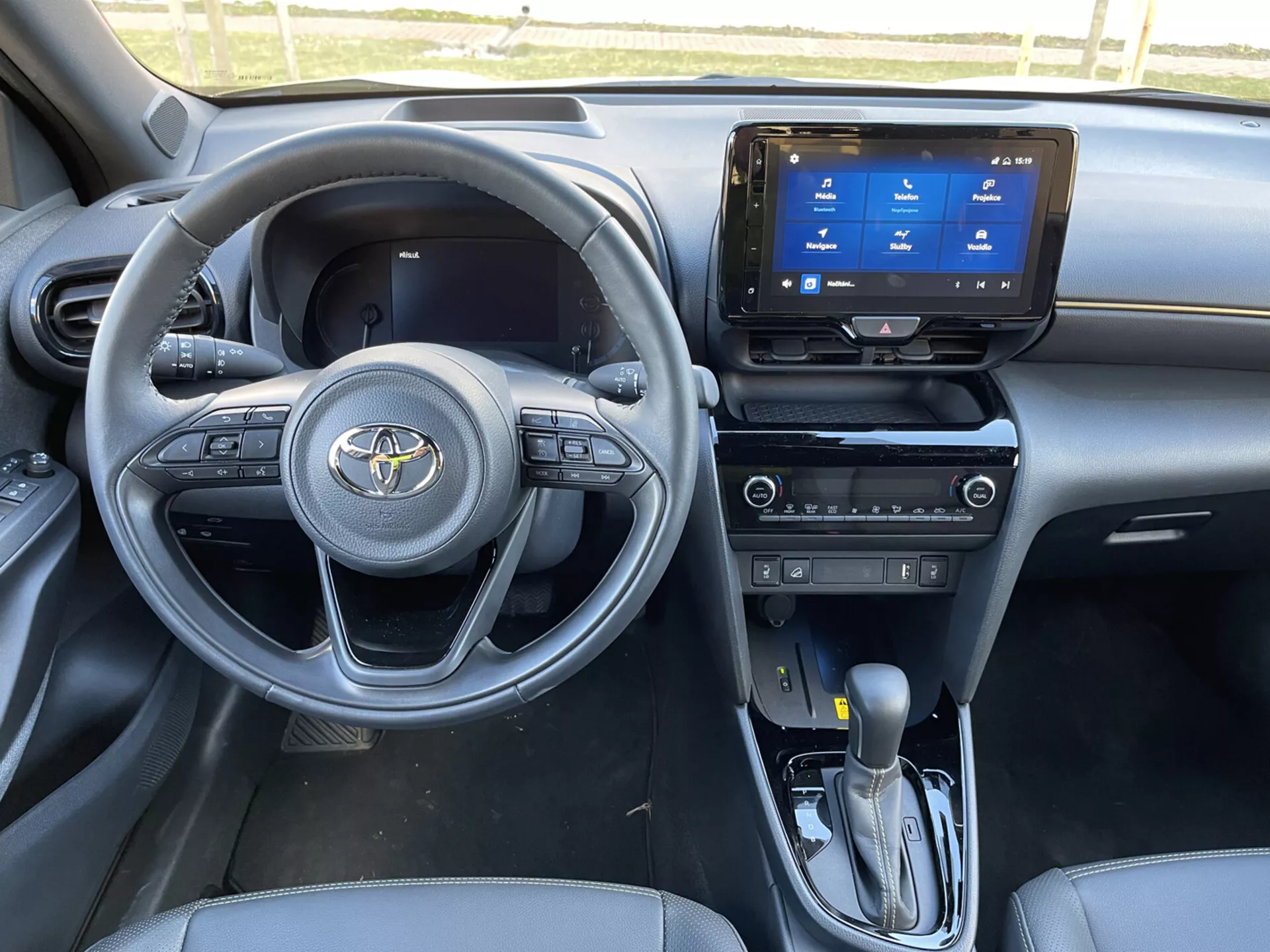 Notranji pogled na Toyota Yaris-Cross 2021 s poudarkom na voznikovem kokpitu z večfunkcijskim tristopenjskim volanom, digitalno armaturno ploščo, zaslonom na dotik z informacijsko-zabavnim sistemom s funkcijami povezljivosti, ročnimi upravljalniki za klimo z dvema conama, avtomatsko prestavno ročico z ročnim načinom, sedeži oblečenimi v material, podoben usnju, kontrolniki električnih oken na vratih voznika in vidnimi držali za kozarce ob prestavni ročici, ki prikazujejo napredno avtomobilsko oblikovanje in sodobne funkcije avtomobila.