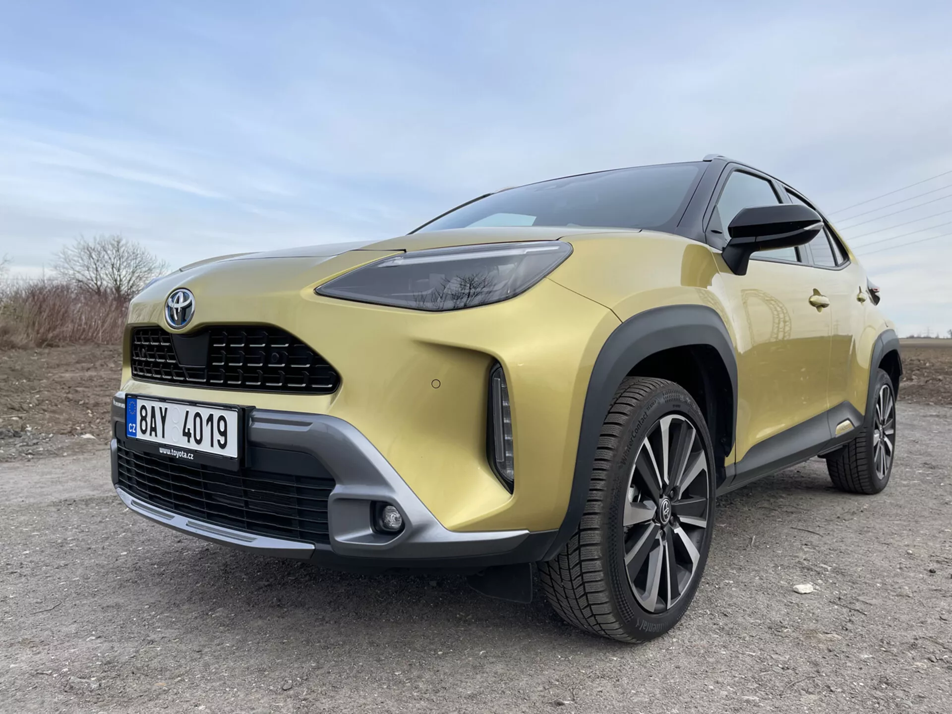SUV Toyota Yaris-Cross 2021 v zlati barvi prikazuje svojo sprednjo levo četrtino iz nizkega širokokotnega pogleda, poudarjajoč LED žaromete, sprednjo masko, odbijač, zlitinsko platišče in moderne oblikovalske elemente na neasfaltirani površini