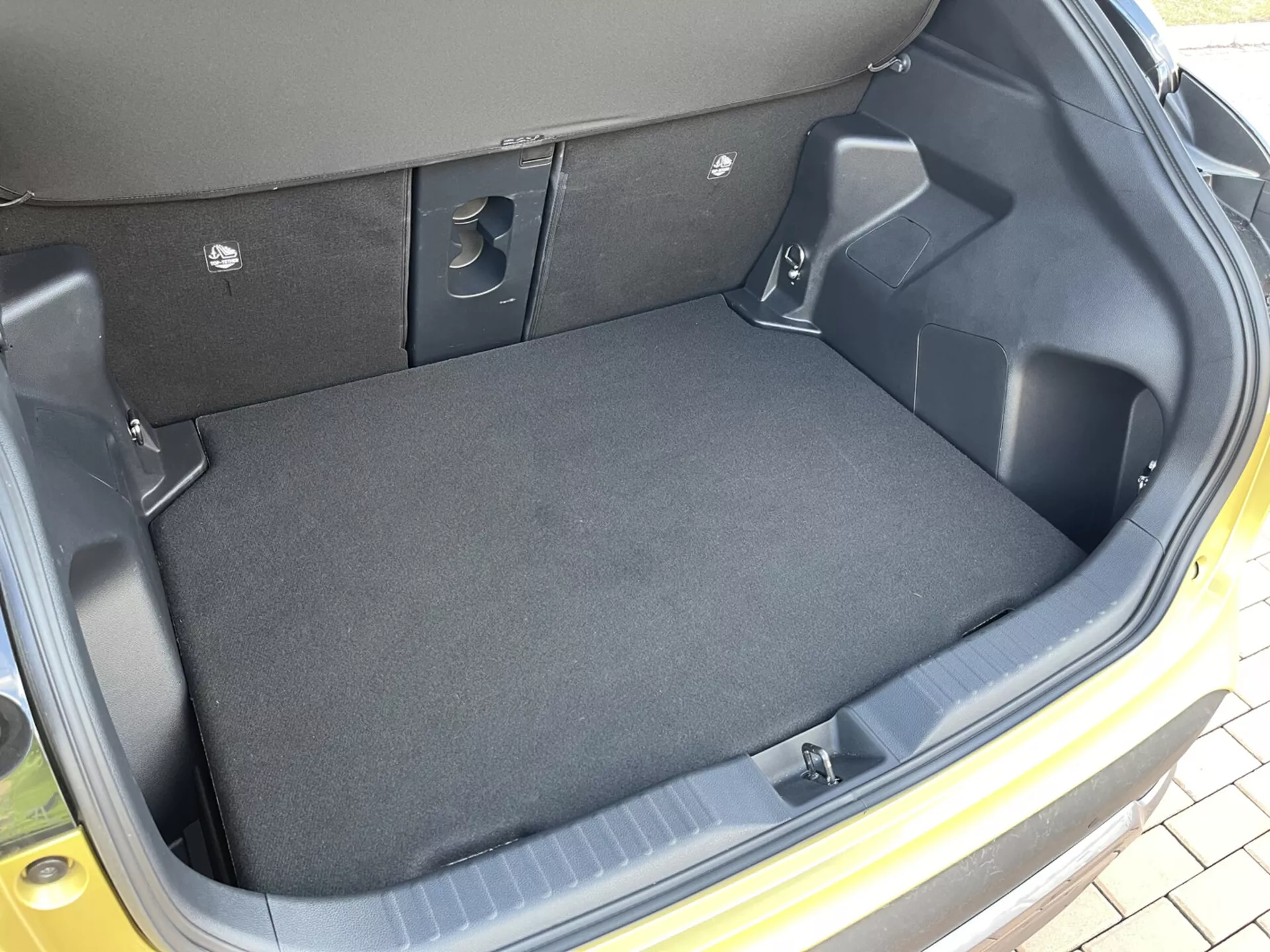 Pogled na notranjost prtljažnika Toyota Yaris Cross 2021, ki prikazuje prazen, s preprogo obložen tovorni prostor z oblikovanimi plastičnimi stranicami, vdolbinami za shranjevanje, izrezi za dostop do opreme in votlinami za zložljive zadnje sedeže