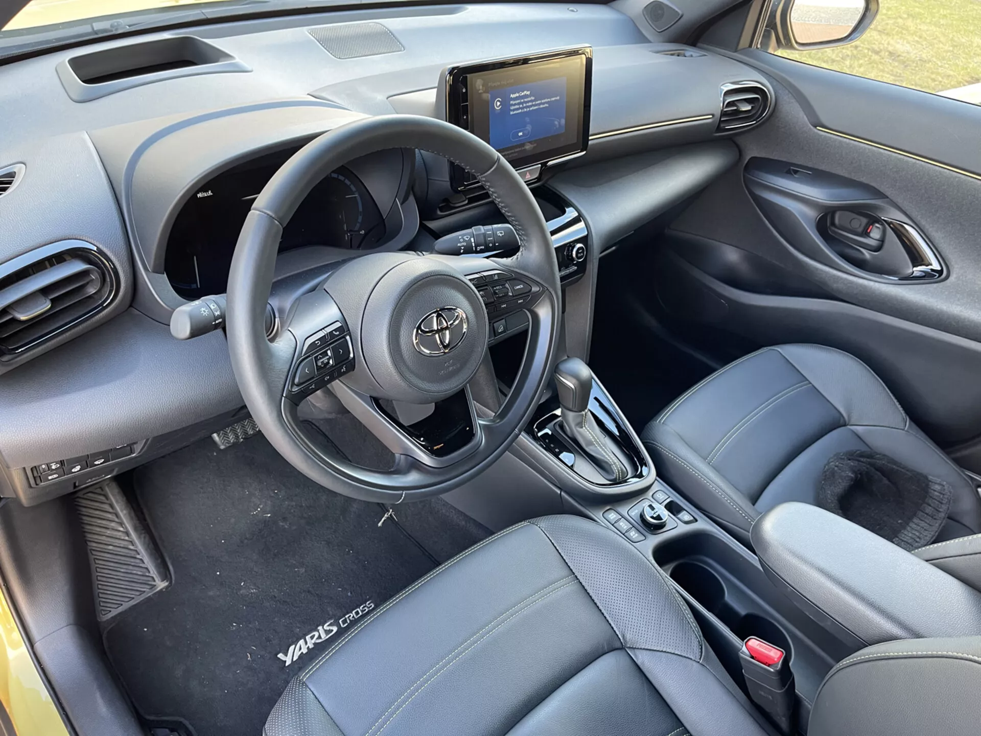 Notranji pogled Toyote Yaris-Cross 2021 iz zadnjih sedežev, poudarja volan z upravljavci, zračnike na armaturni plošči, sredinsko konzolo z infotainment sistemom, upravljavce za klimatsko napravo, ročico avtomatskega menjalnika, prednja vedra sedeža s podobnim usnjem z oznako Yaris Cross, plošče vrat z električnimi upravljavci in moderno, elegantno obliko kabine.