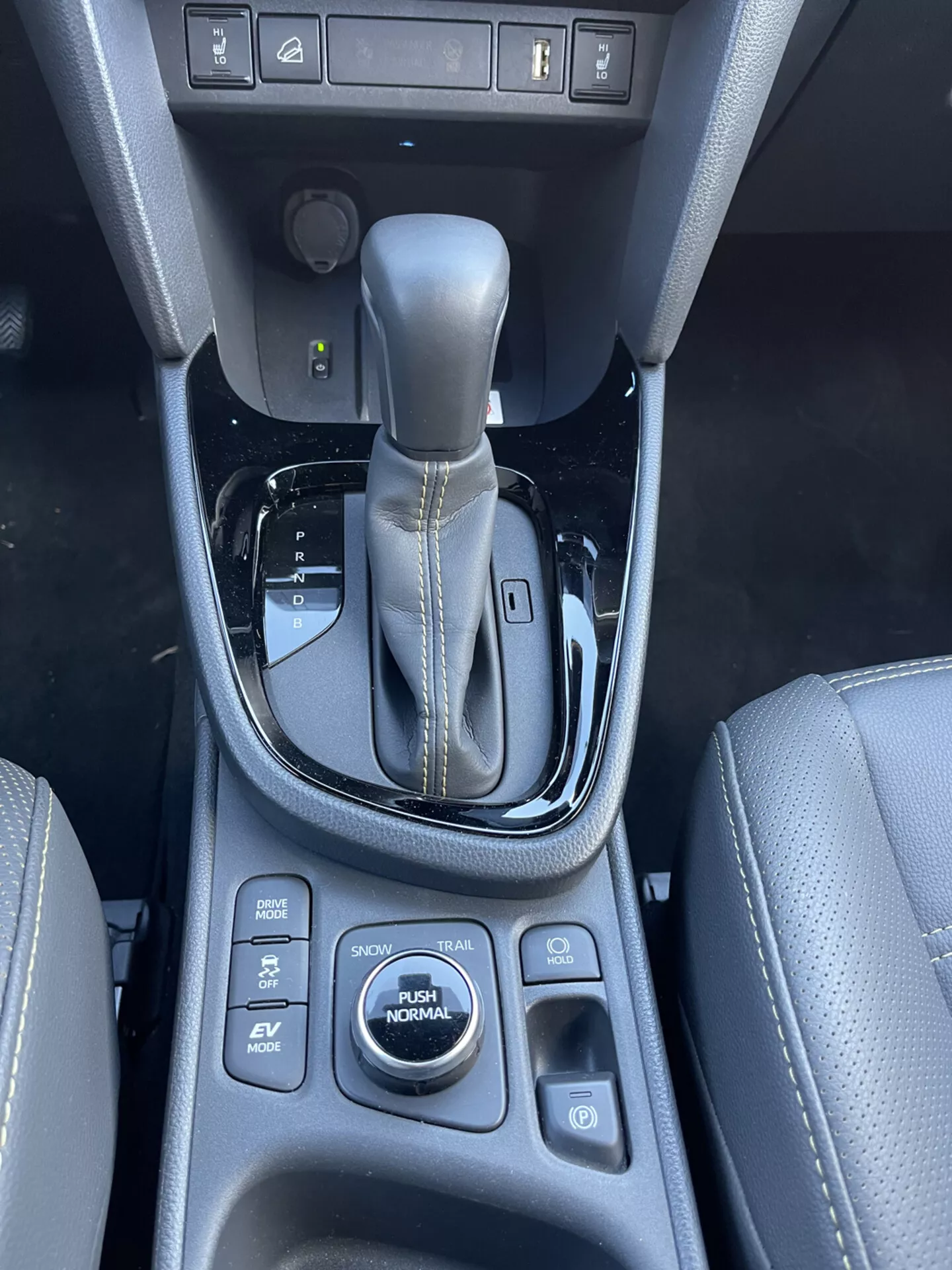 Notranja sredinska konzola Toyota Yaris Cross 2021 z avtomatsko prestavno ročico v položaju parkiranja, obkrožena z nadzornimi gumbi za pomoč pri parkiranju, zadrževanje zavore, parkirne senzorje in načine vožnje, vključno z SPORT, ECO, EV, AUTO in NORMAL, z materialom, ki spominja na usnje z detajli šivanja in sijočimi črnimi detajli