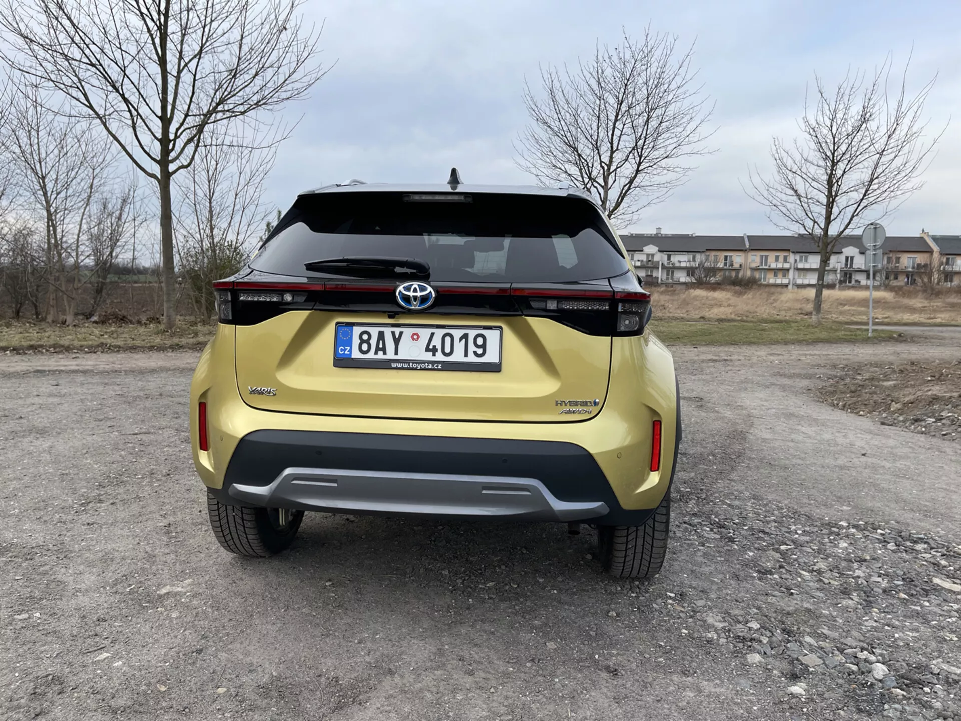 Zadnji pogled na rumeno Toyota Yaris Cross 2021, posnet na višini oči, ki prikazuje njeno sodobno oblikovanje, horizontalno sestavo zadnjih luči, emblema Toyota, ime modela Urban Cruiser, registrska tablica, zadnji odbijač kontrastnega dizajna z integriranimi parkirnimi senzorji in ročaj prtljažnika na predmestnem ozadju.
