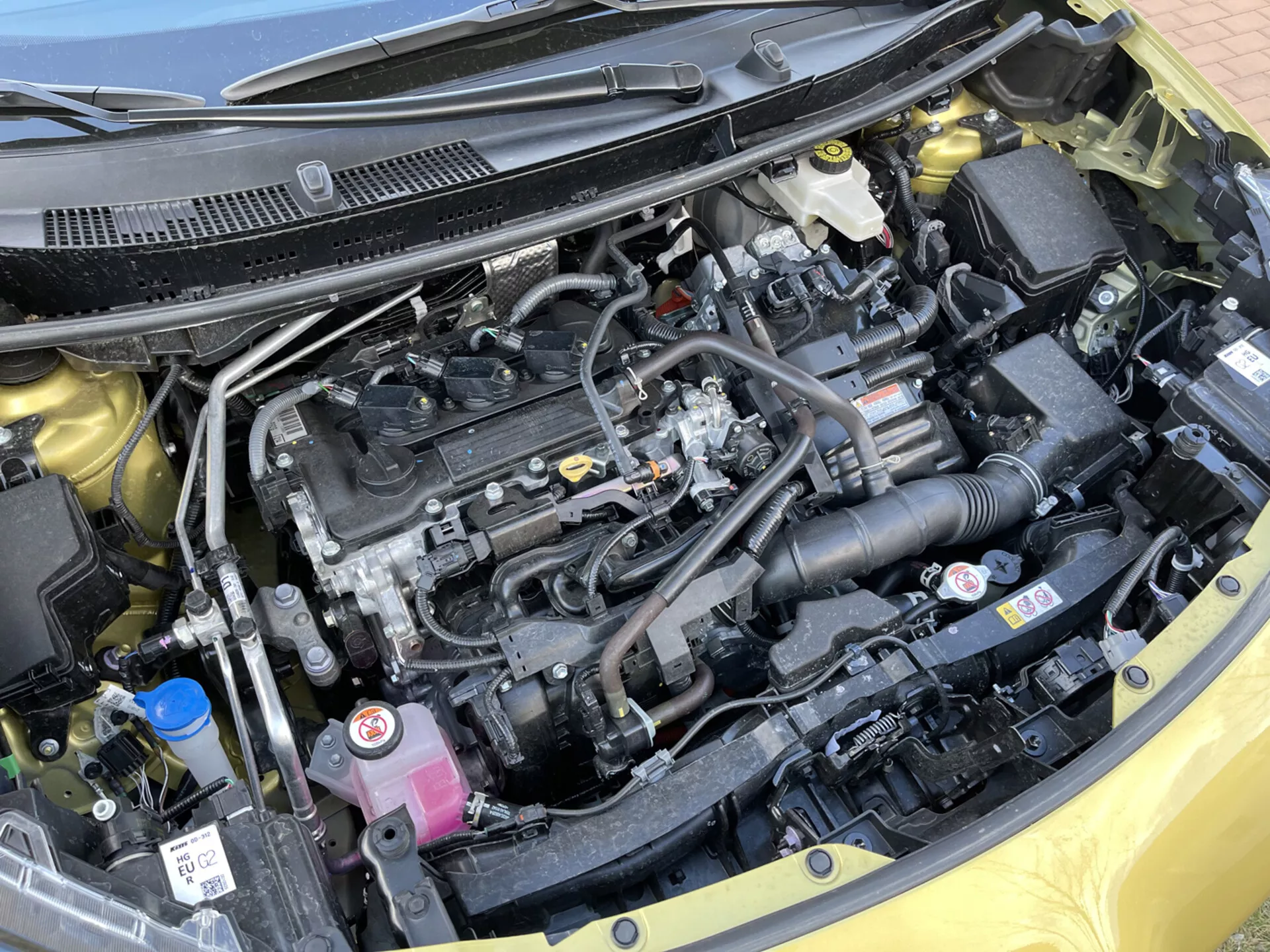 Podroben pogled na motor Toyota Yaris-Cross 2021, ki prikazuje vrstni motor, črni plastični pokrov motorja, sistem za dovod zraka, rezervoarji za hladilno in zavorno tekočino, električne komponente, baterijo, radiator in urejeno postavitev z varnostnimi nalepkami.